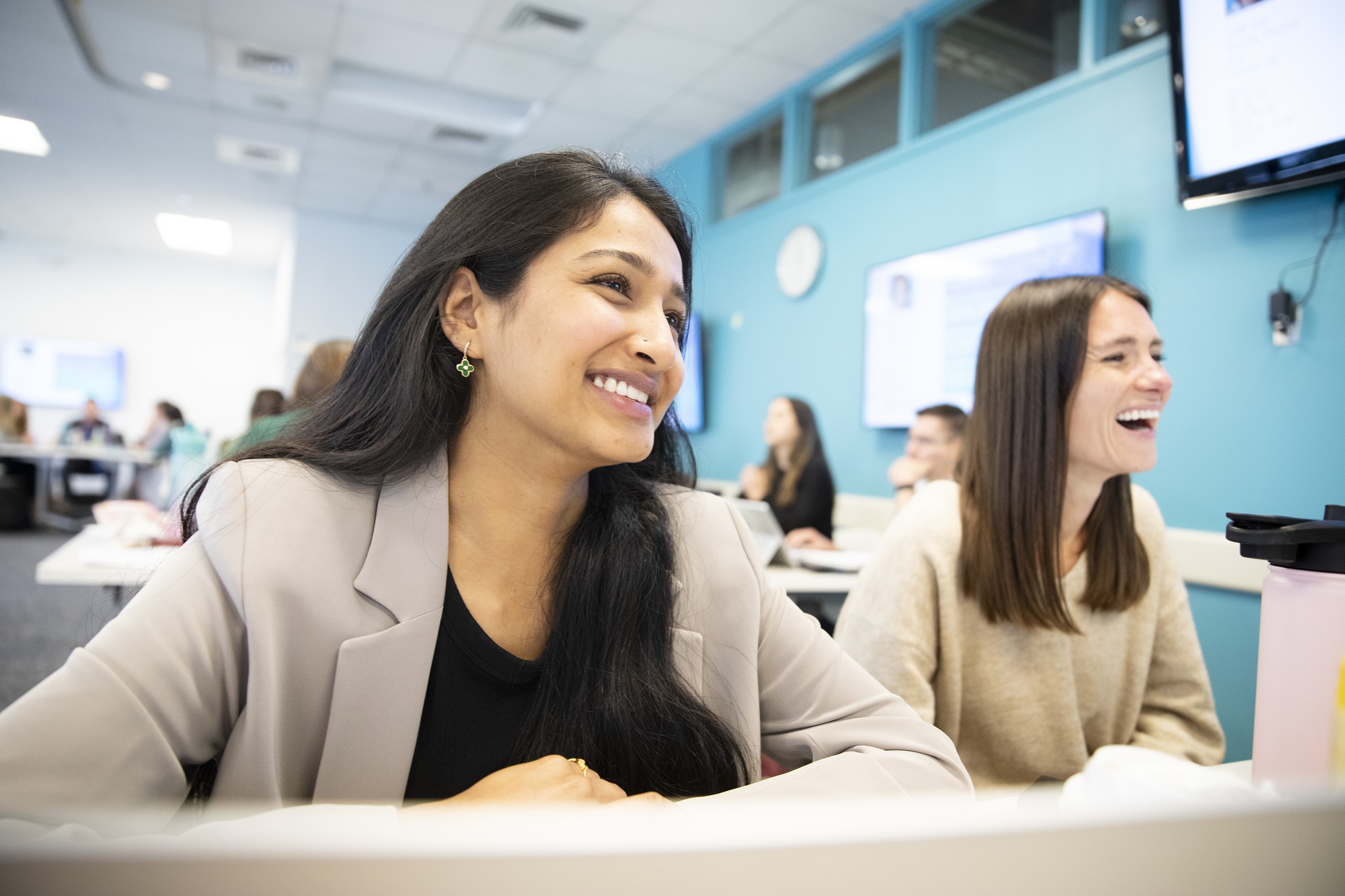 two females sit at a desk and laugh at something off camera