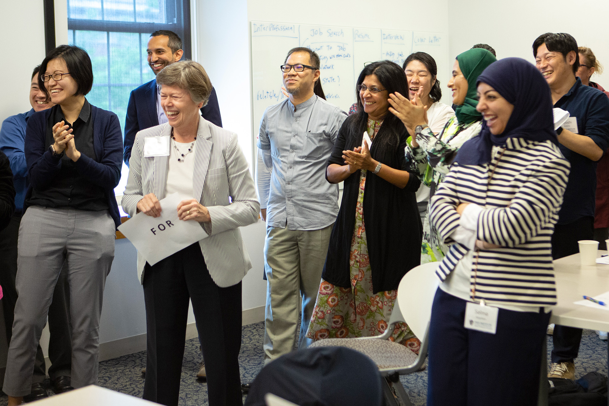 a group of men and women, some Asian and some in hijabs smile and laugh in a classroom setting