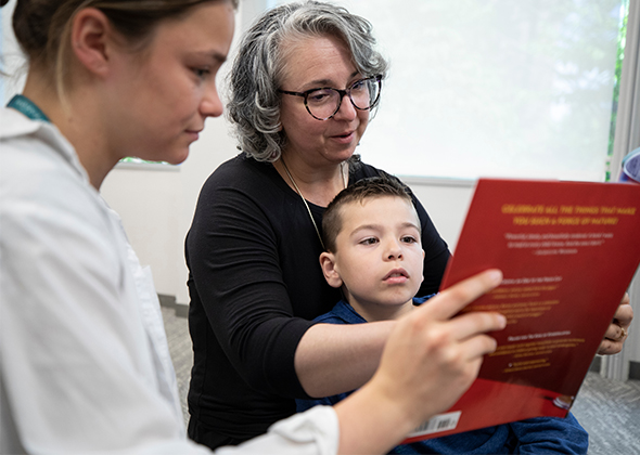 woman reads to boy while woman in lab coat helps hold the book