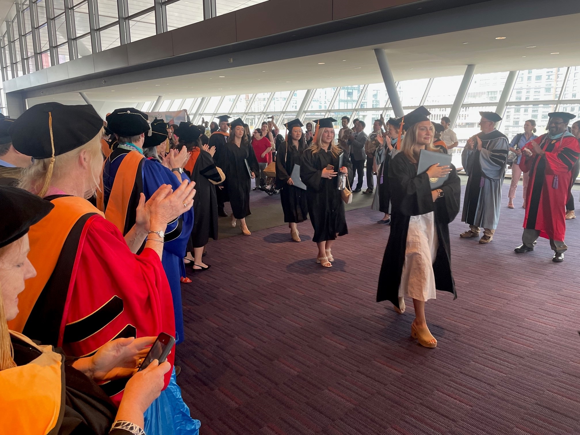 people in colorful graduation regalia clap as people wearing black caps and gowns walk in a line