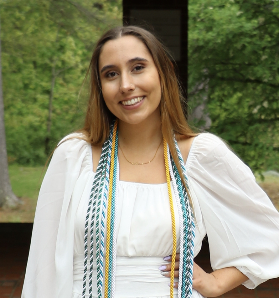 Maegan Mannes is in a white dress with graduation cords and has long brown hair
