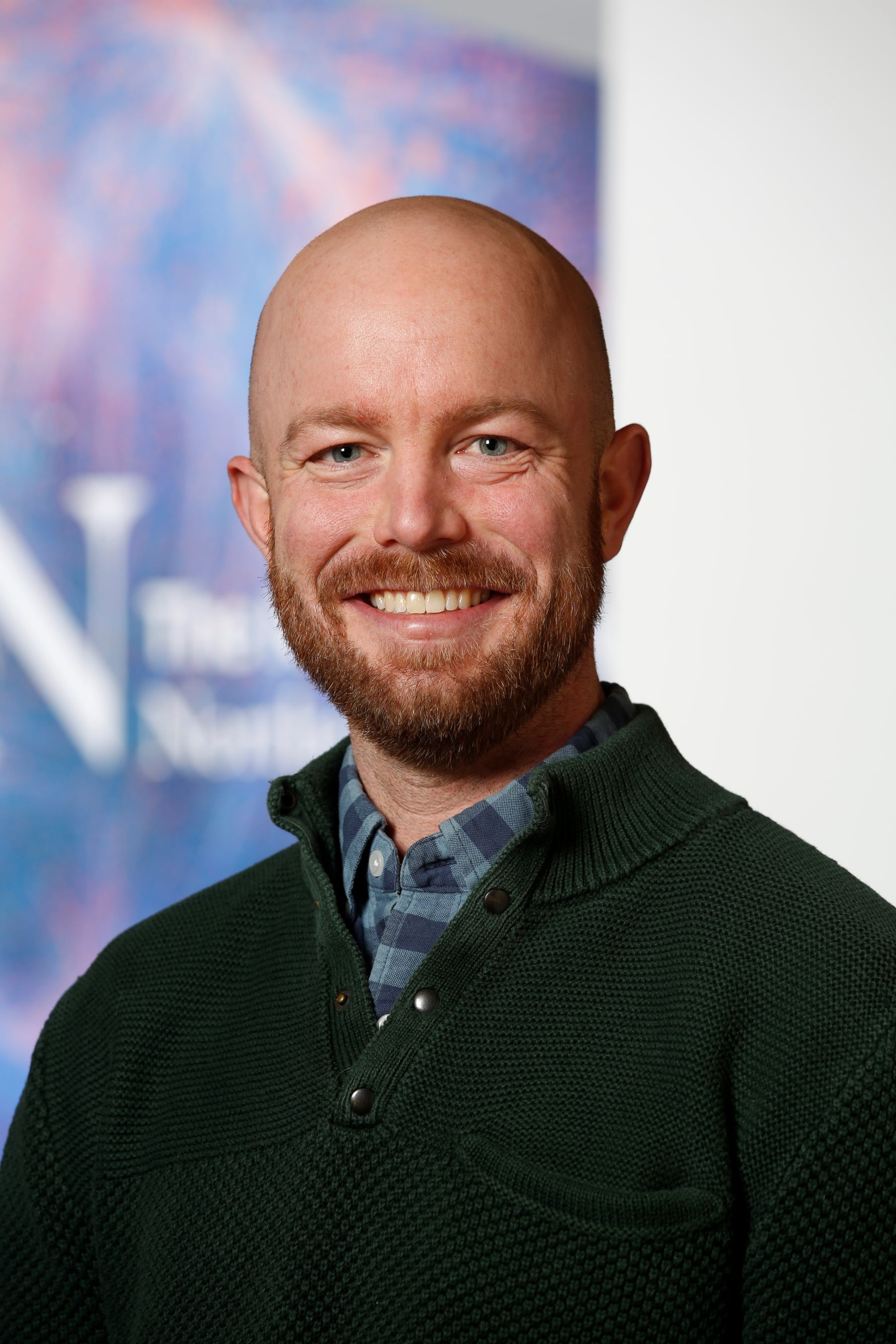 Robert Cavanaugh has a bald head and a brown beard. He is wearing a black sweater over a blue checkered dress shirt.