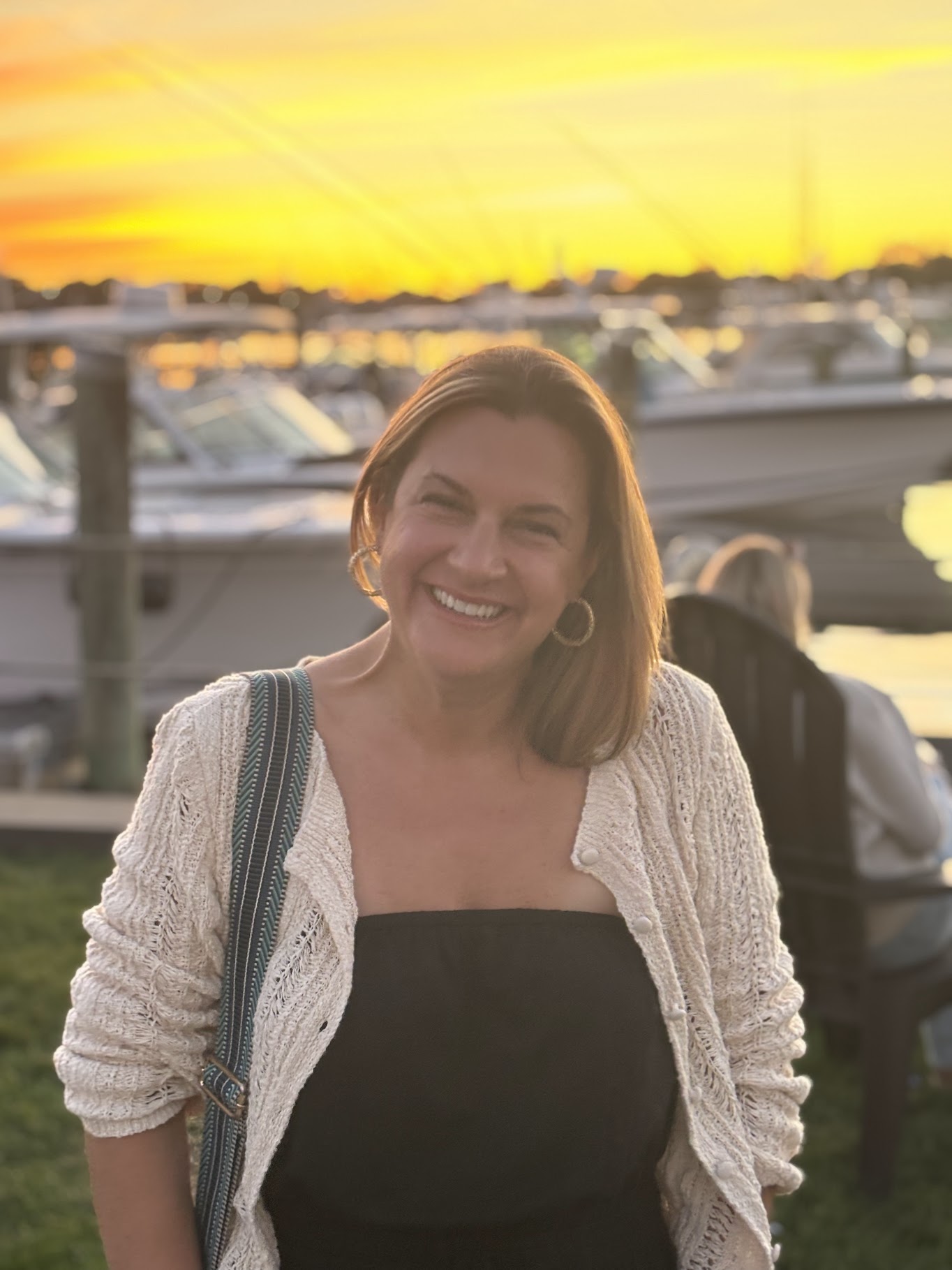 Woman wearing a black shirt and white sweater smiling and looking at camera. There is a sunset and boats on the water in the background.