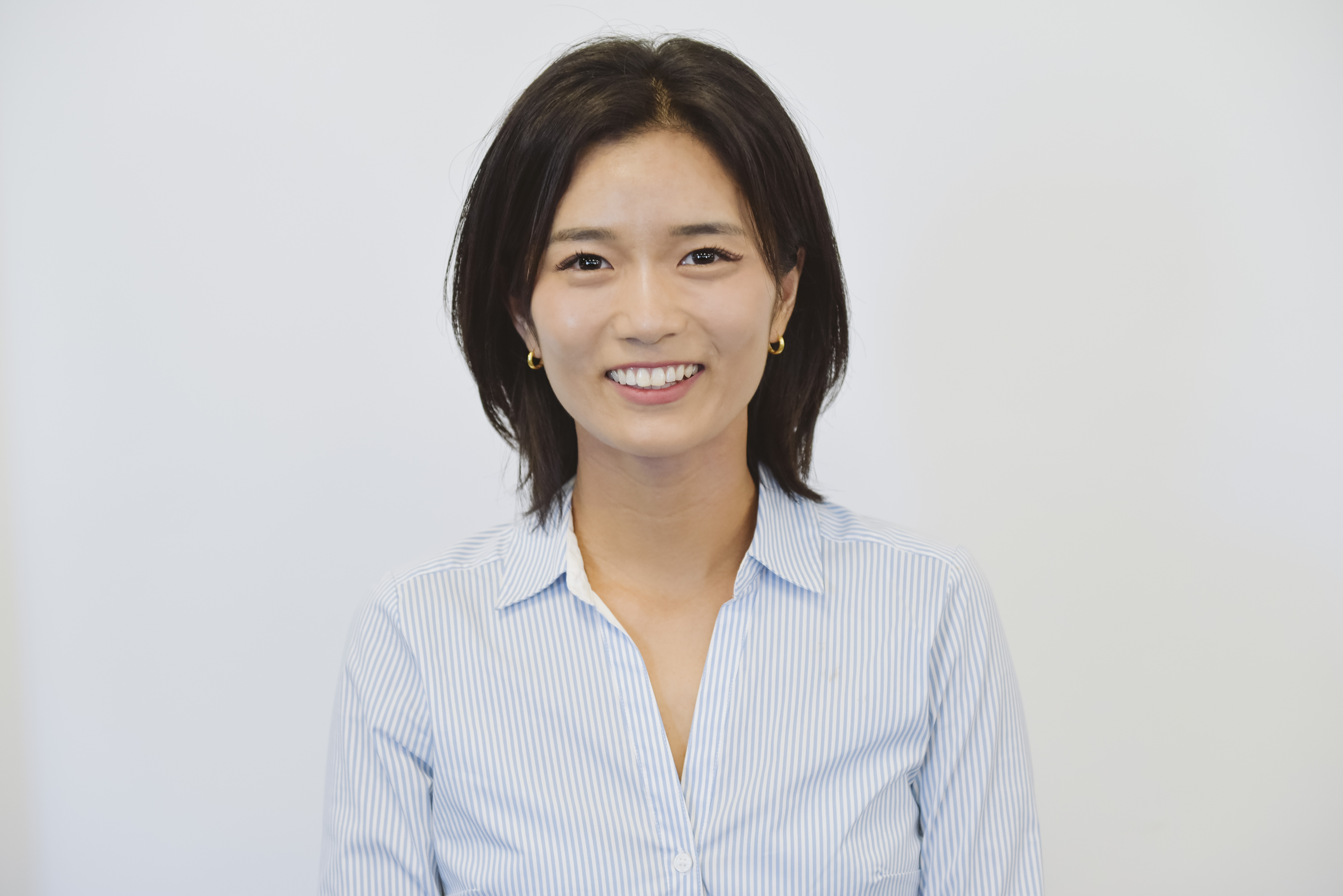 Headshot of woman with black hair standing against a solid white background wearing a blue collared shirt