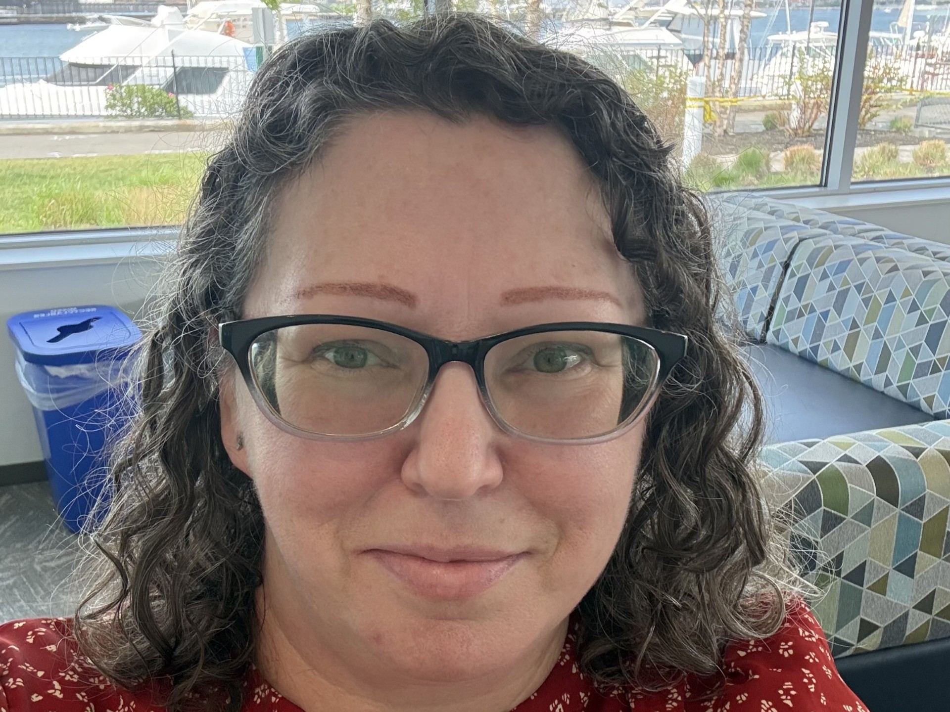 Head shot of Woman with curly dark hair wearing glasses and a red shirt in front of a waterfront background.