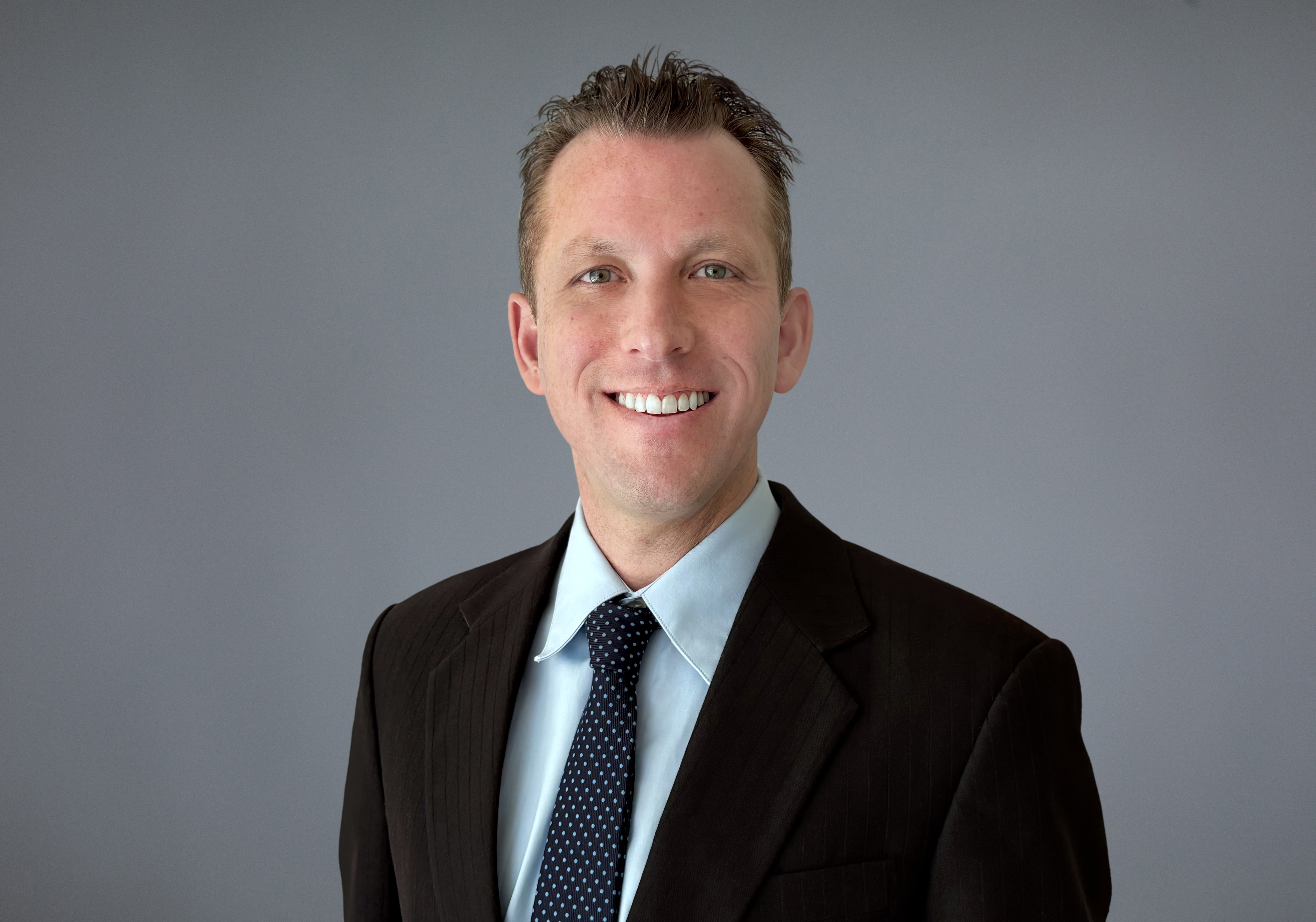 Headshot of Jason Richter standing against a solid gray background, wearing a blue collared shirt, a black blazer and, and blue necktie with white dots