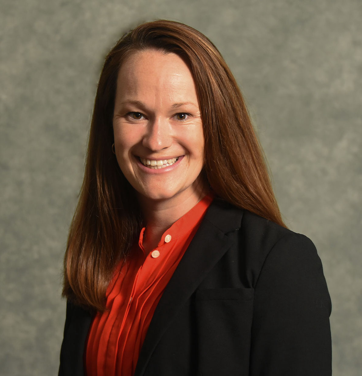 Headshot of Adrian Wright-FitzGerald with long dark blond hair, wearing an orange shirt and a black jacket against a gray background
