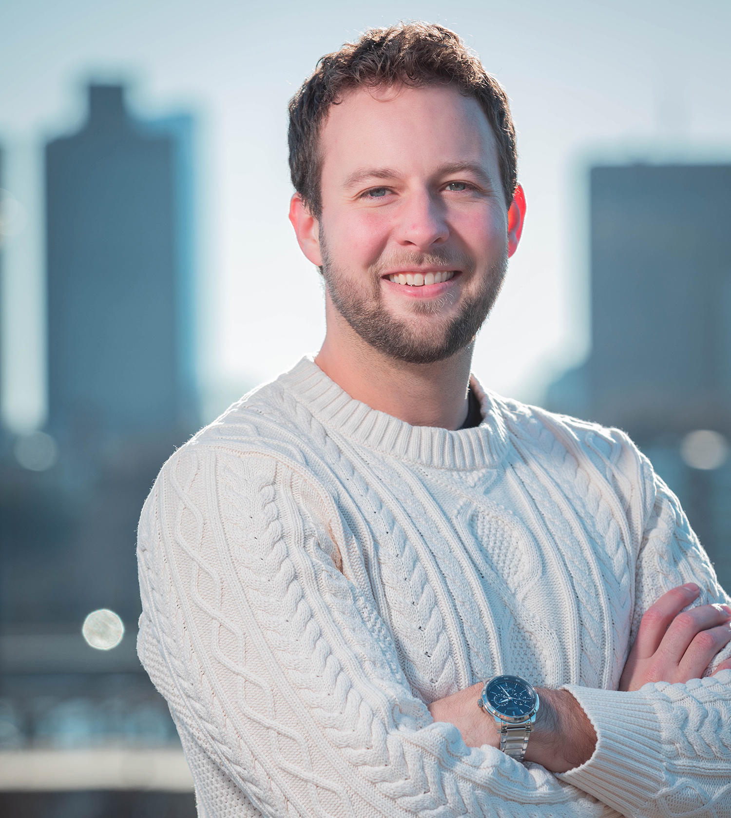 Headshot of Ian Karby standing with his arms crossed against a city skyline background wearing a white sweater and a watch.