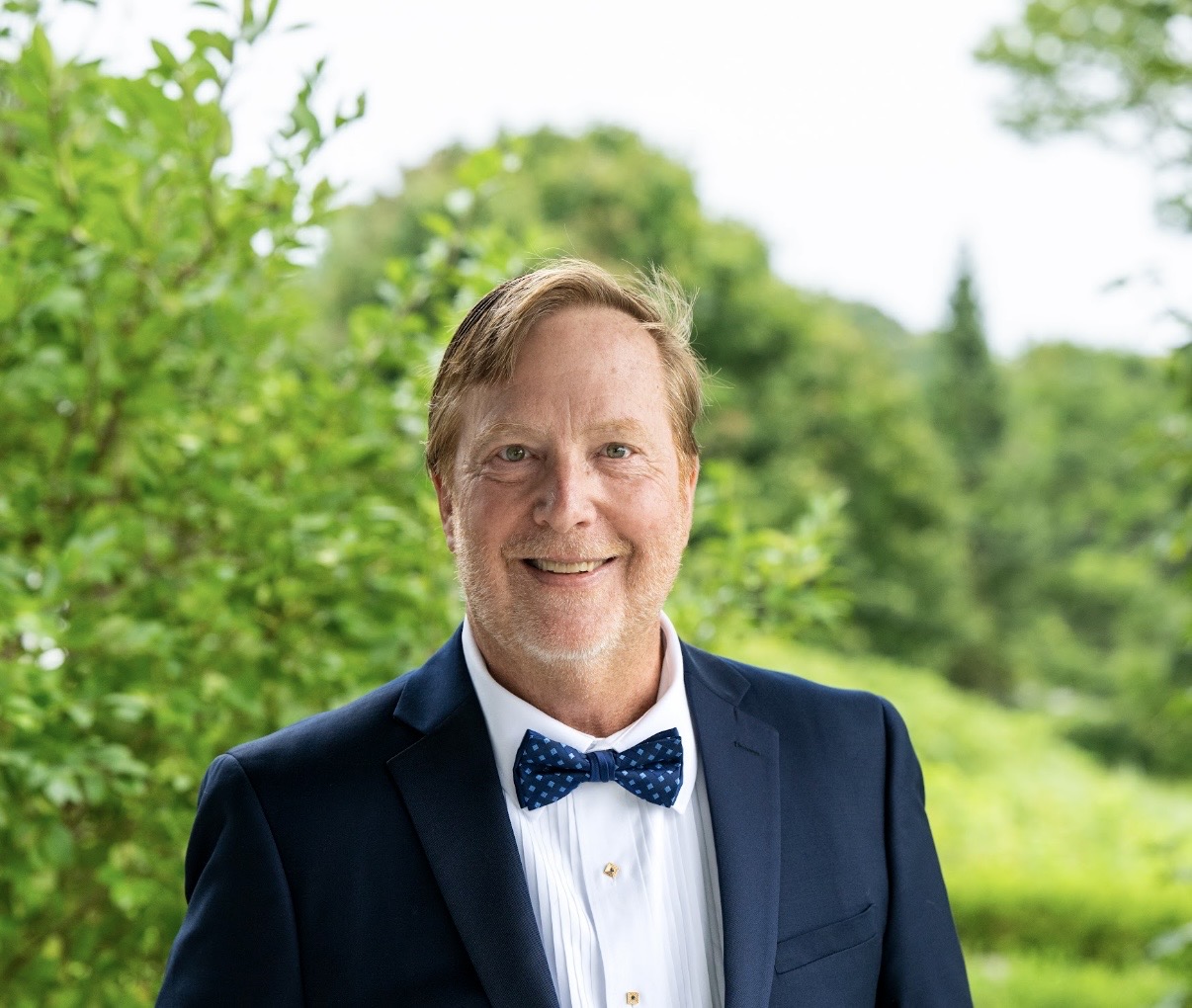 Headshot of Scott Goodspeed wearing a white button down shirt, dark blue blazer, and blue bowtie. He is standing in front of a background of green grass and trees