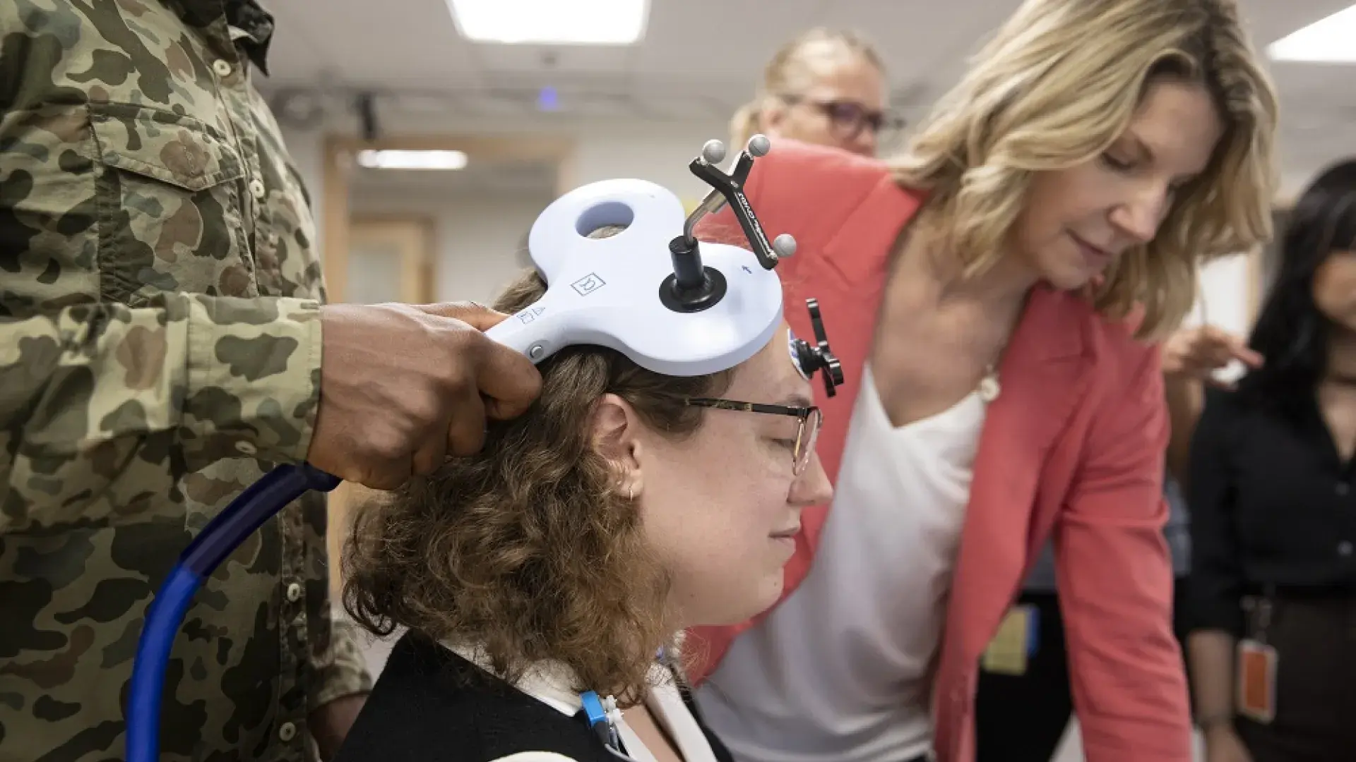 man holds device that looks like a big T to a woman's head while others look at a computer