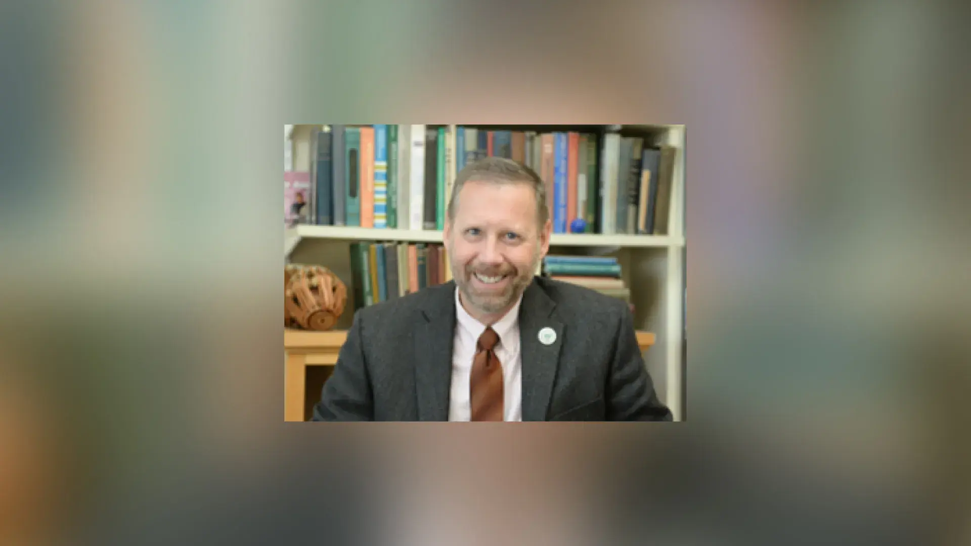 Greg sits in front of a bookcase and wears a grey suit with a maroon tie
