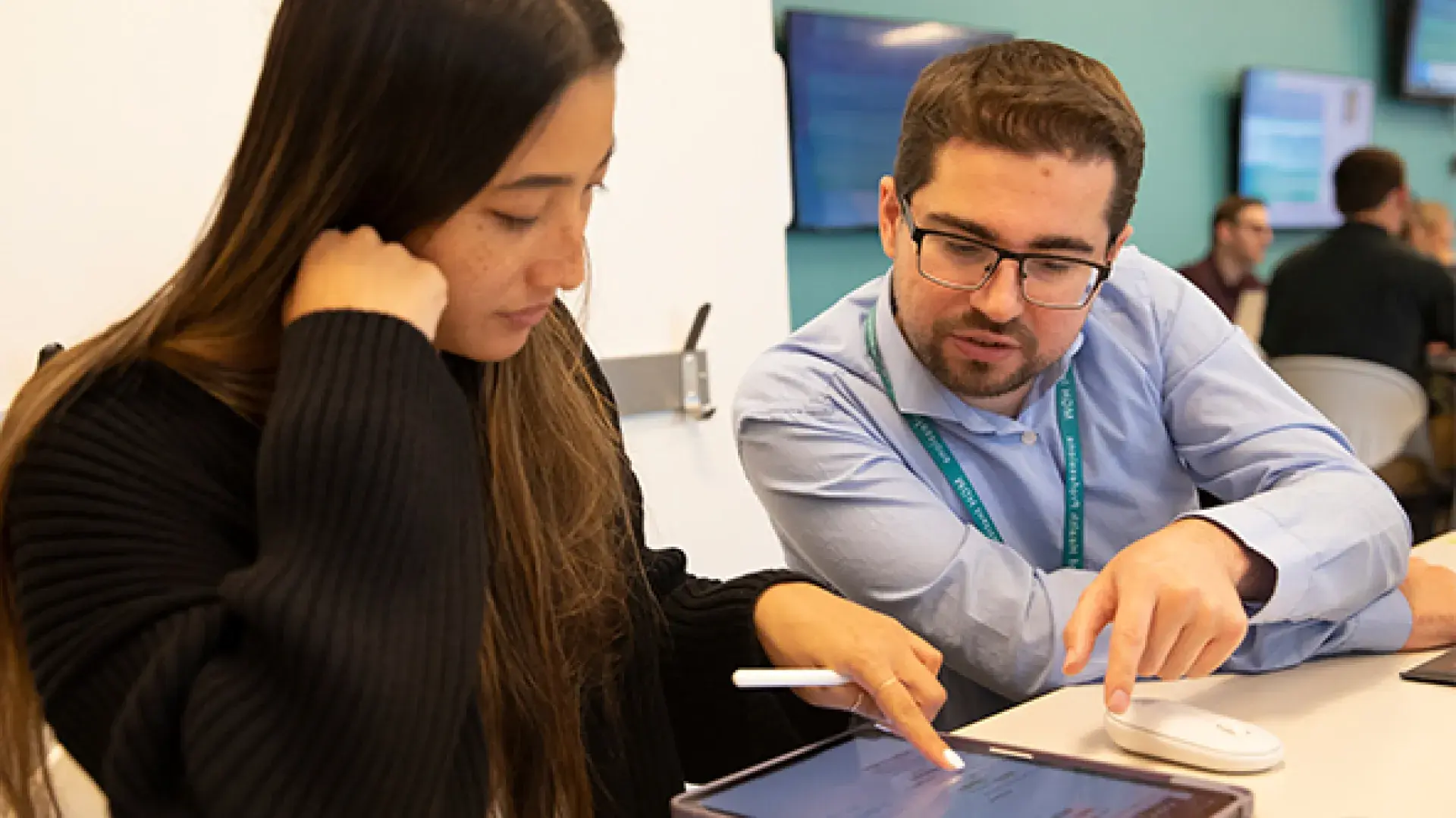 Woman with long hair points at a tablet while a man in a blue shirt looks on
