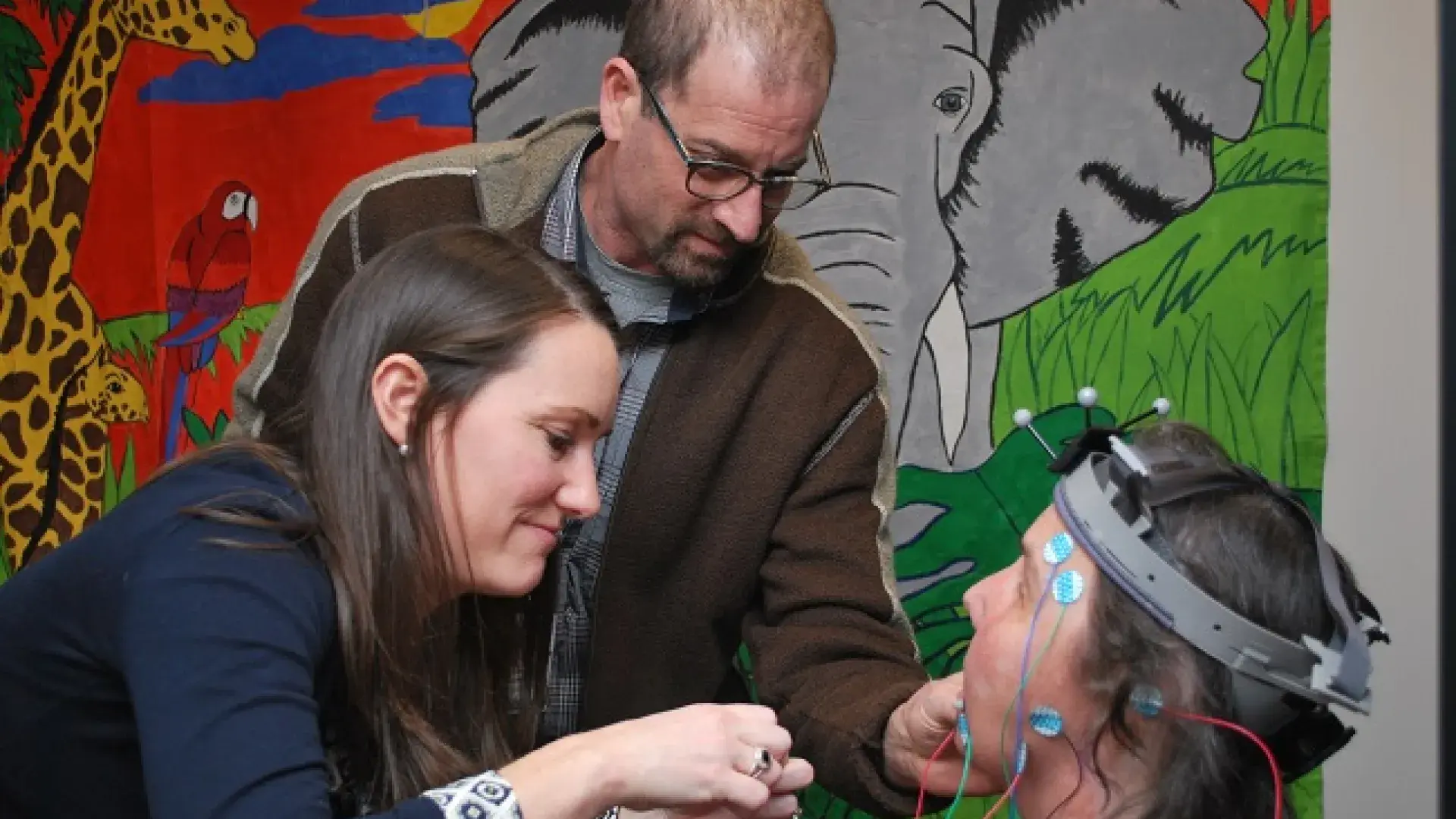 Two SFD Lab Researchers attach sensors to the head of a patient in front of a colorful painting of animals
