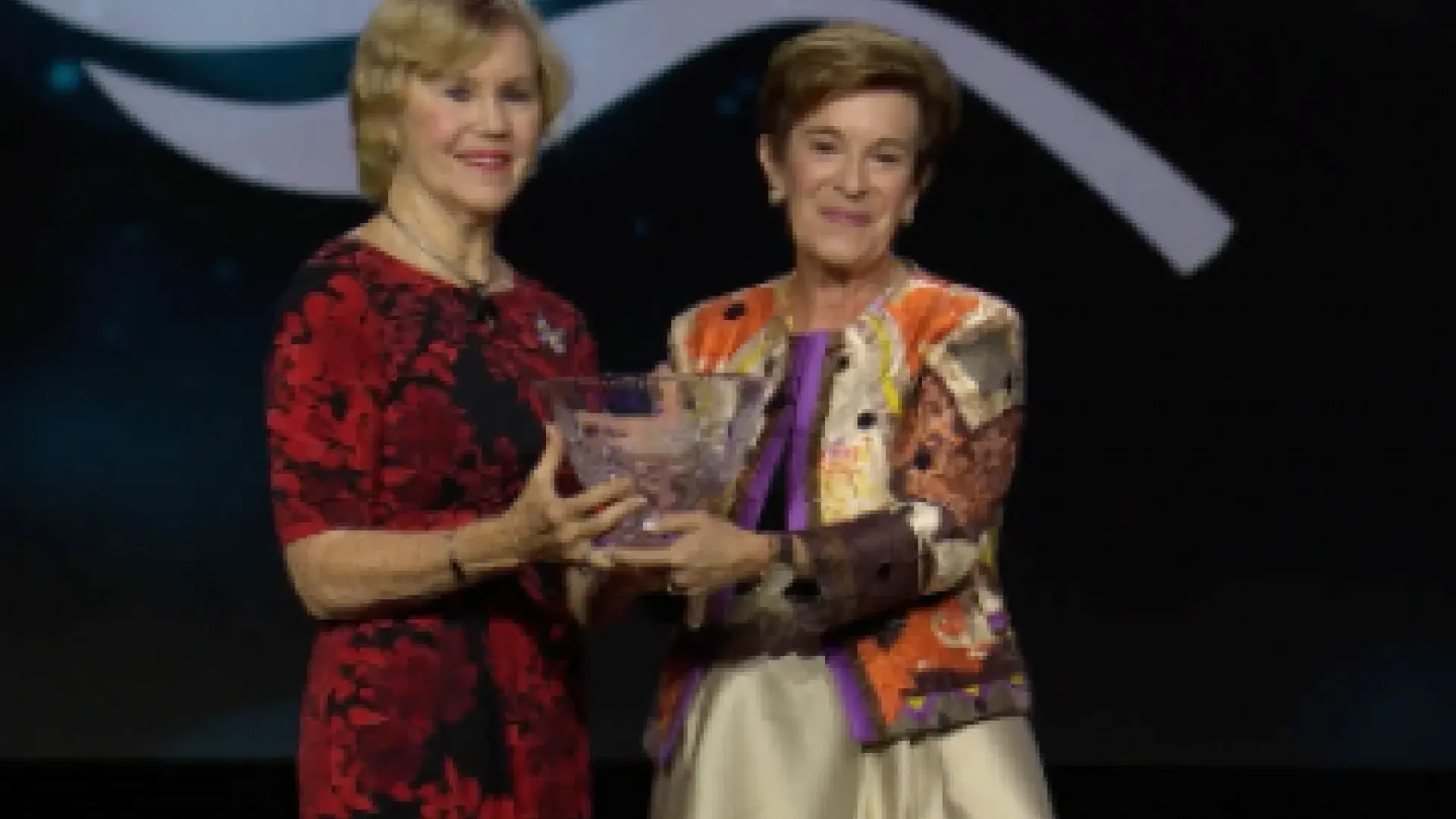 Two women smile on stage and hold a glass award