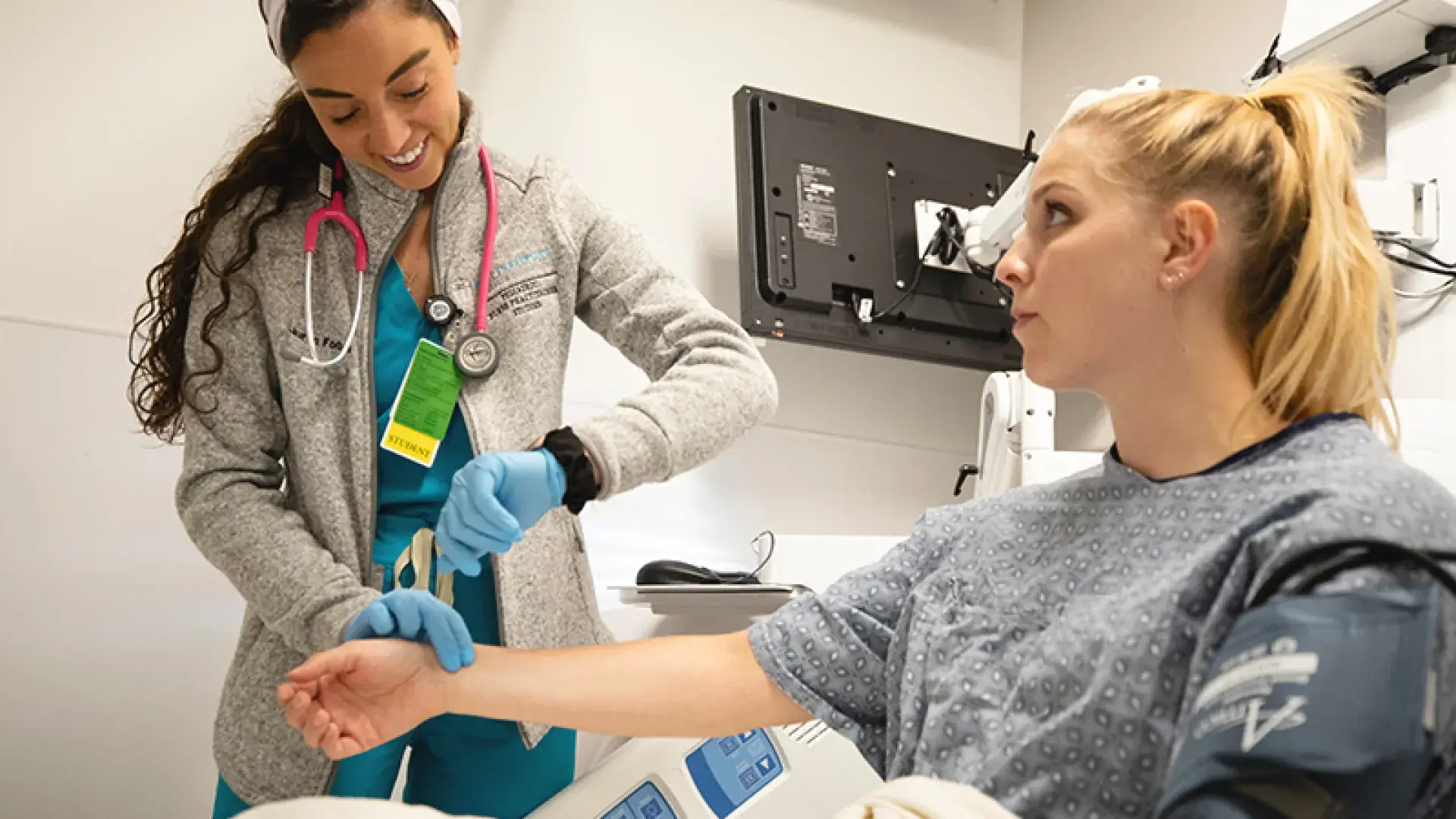 nursing student takes the pulse of another young woman in a hospital gown