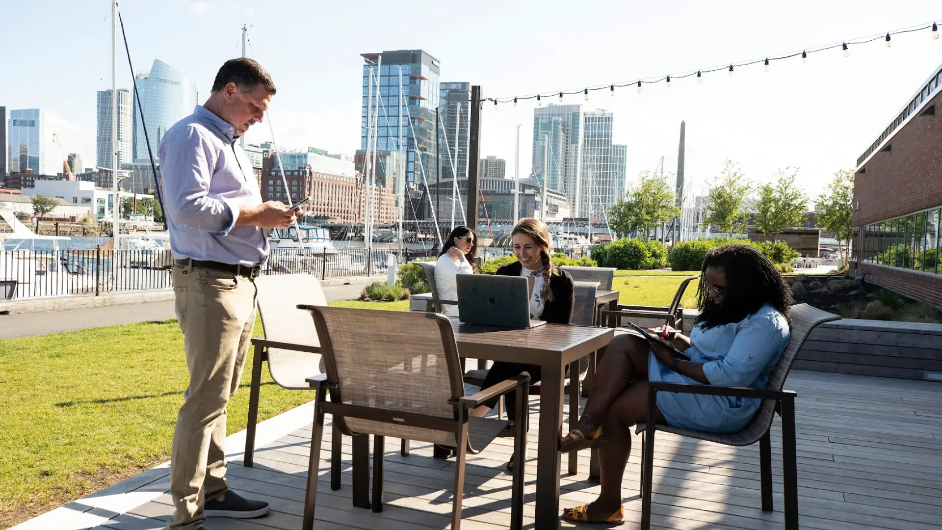 4 people on devices sit outside on a patio with the boston skyline behind them