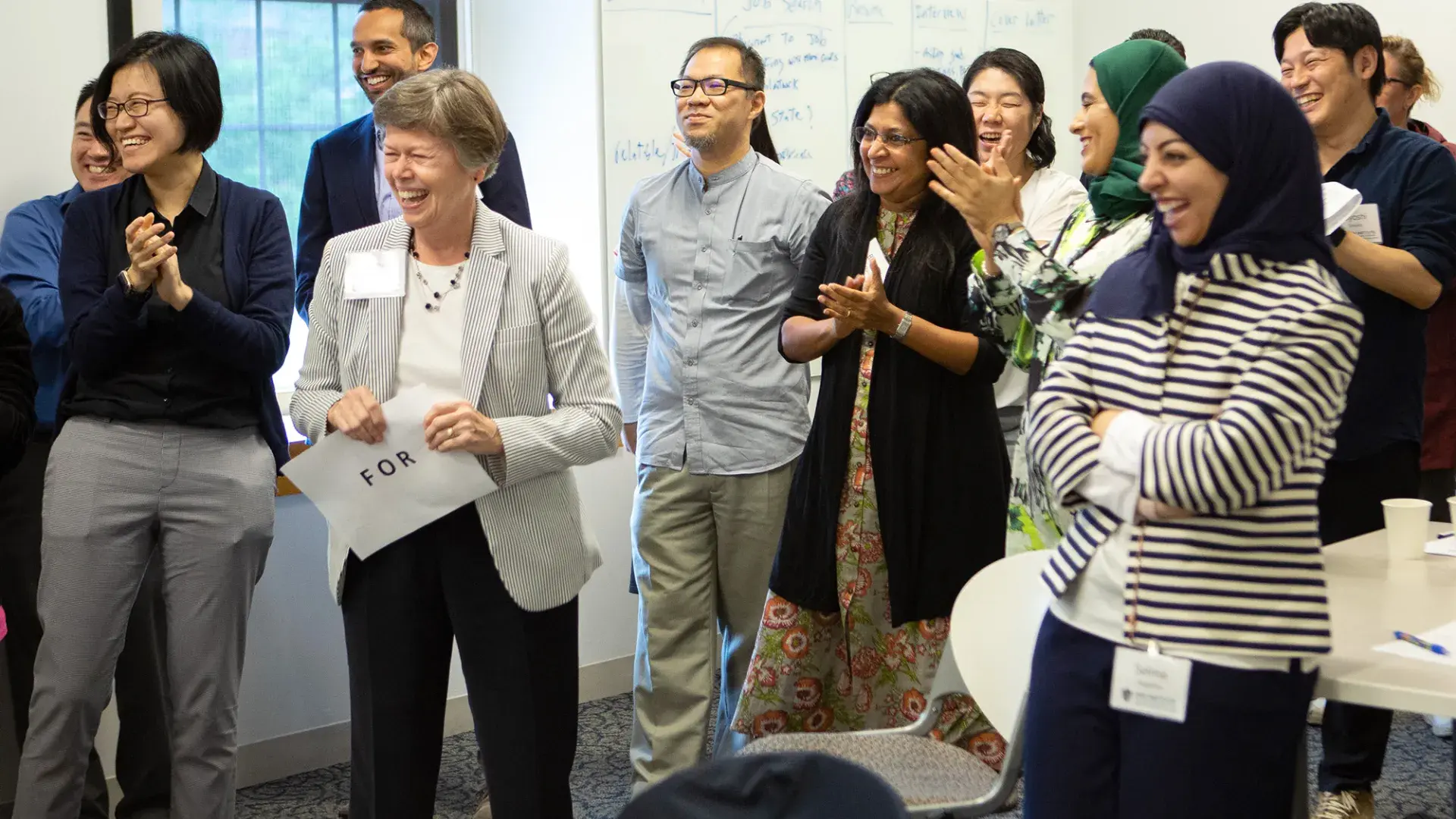 a group of men and women, some Asian and some in hijabs smile and laugh in a classroom setting