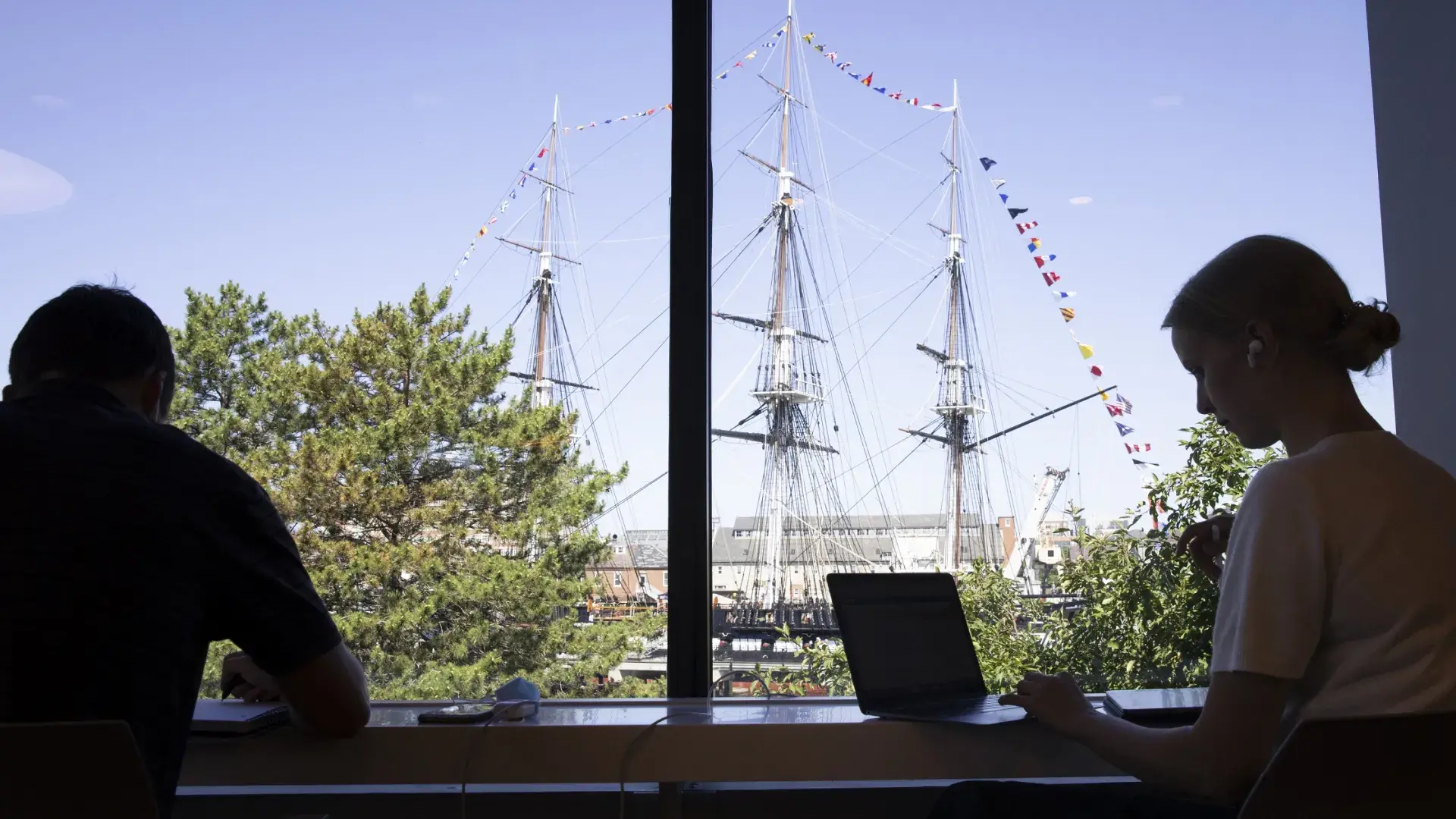 Silhouette of 2 people on laptops in the school in front of a sailing ship outside of the window.
