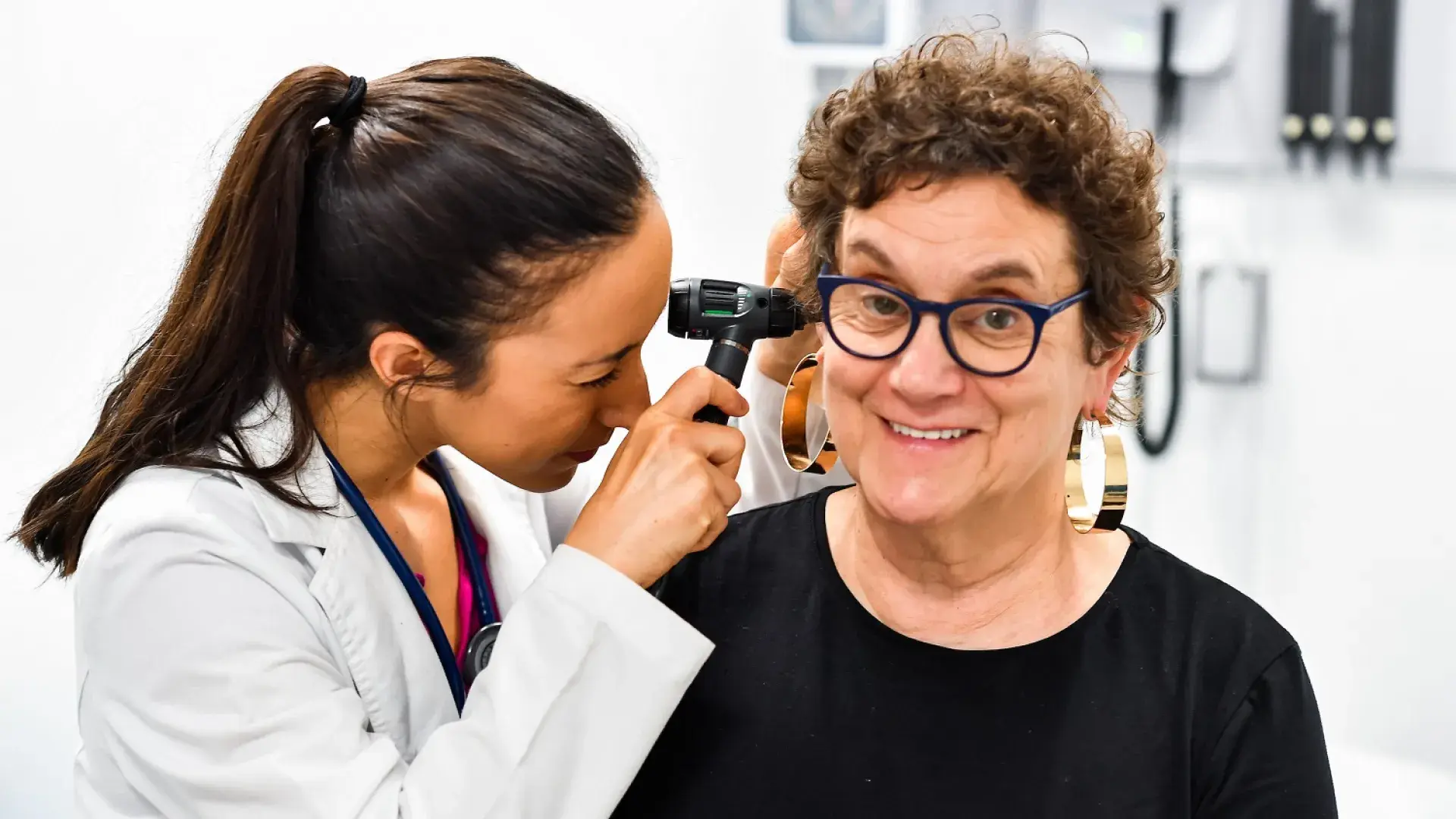 woman in white medical coat looks in the ear of an older woman