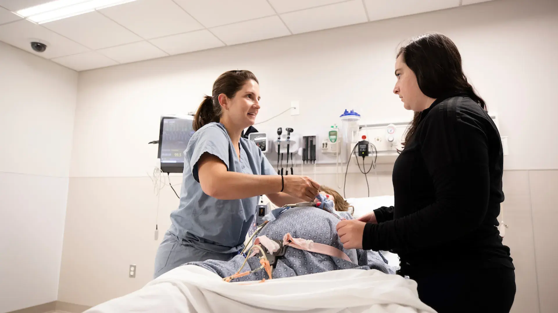 woman in scrubs shows another woman how to adjust a strap on a manikin in a hospital gown