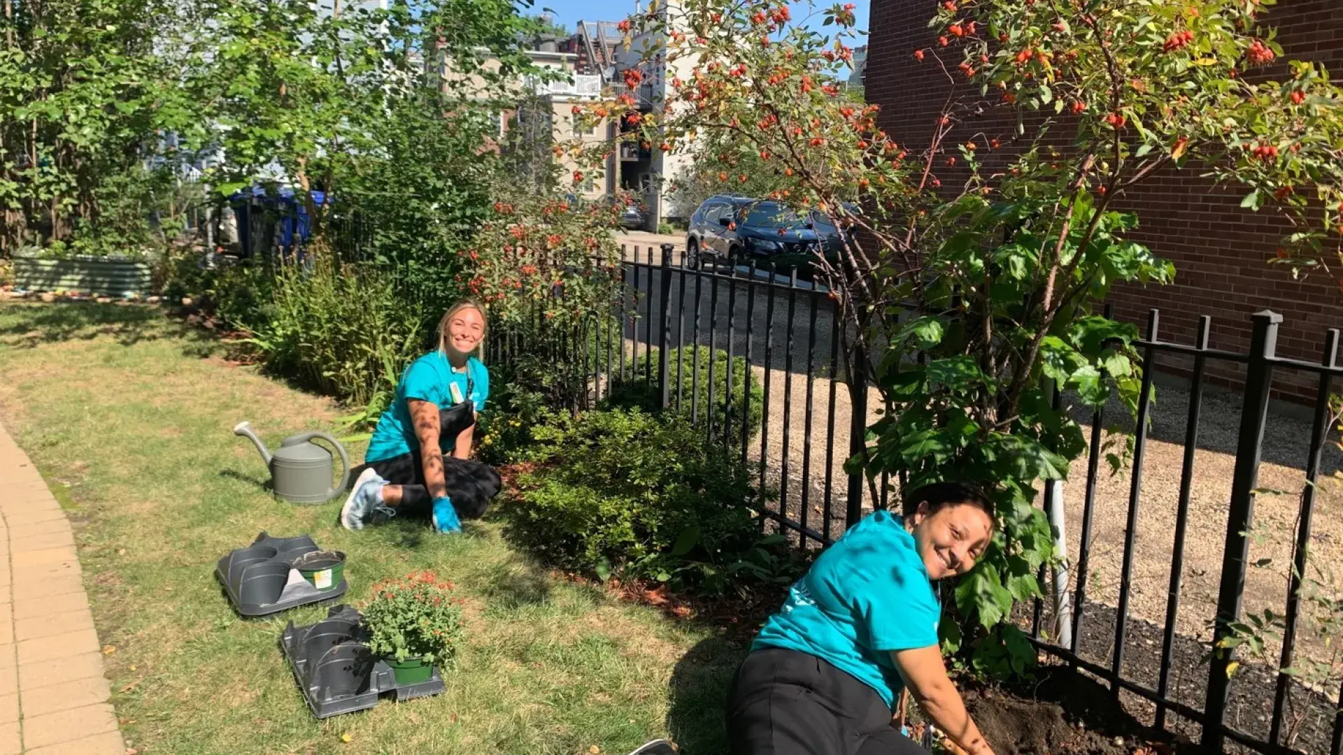 two students sit on the grass they are in the middle of planting flowers along a black fence