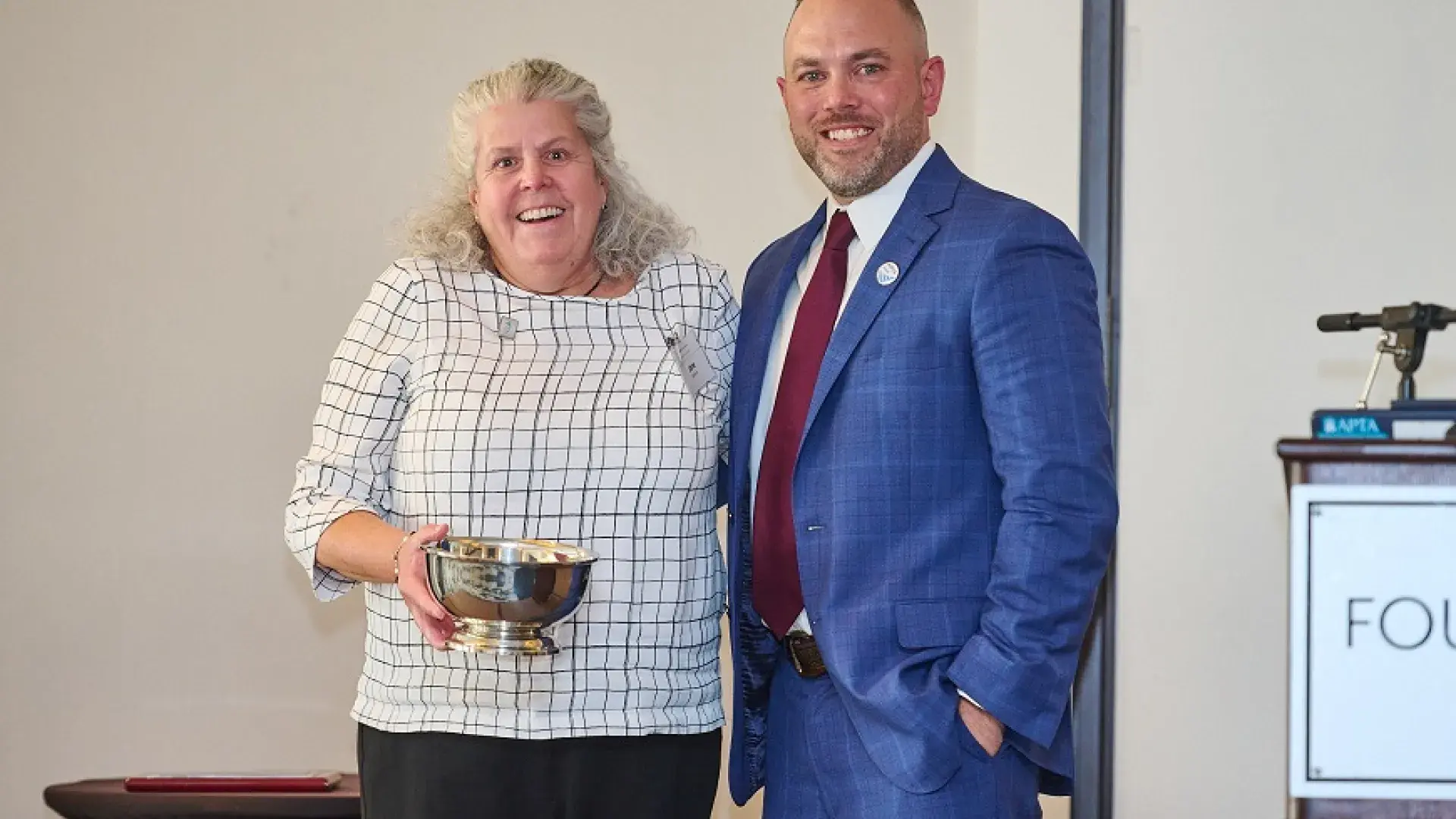 jane smiles and holds a silver bowl and stands with a man in a blue suit