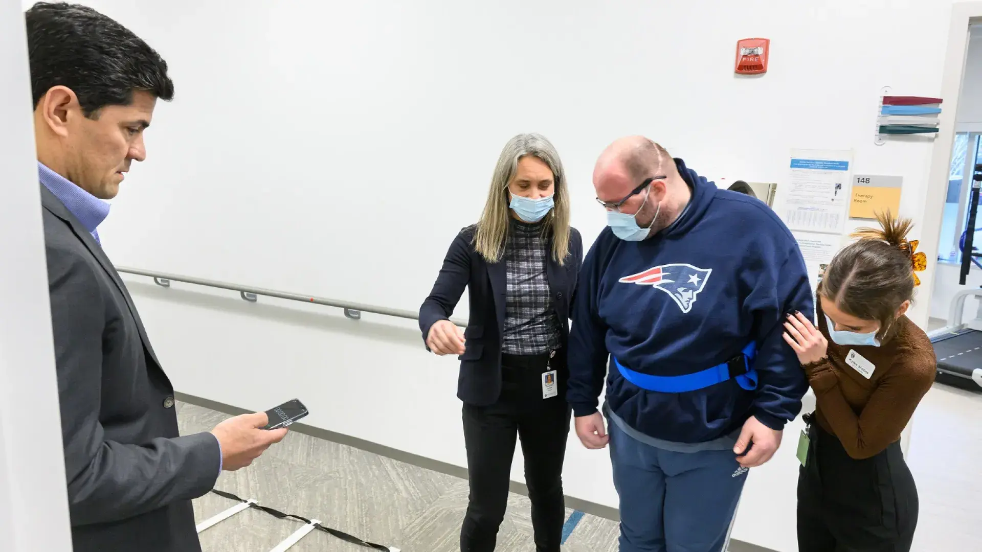 tedy holds a stopwatch while two women help a man wearing a belt