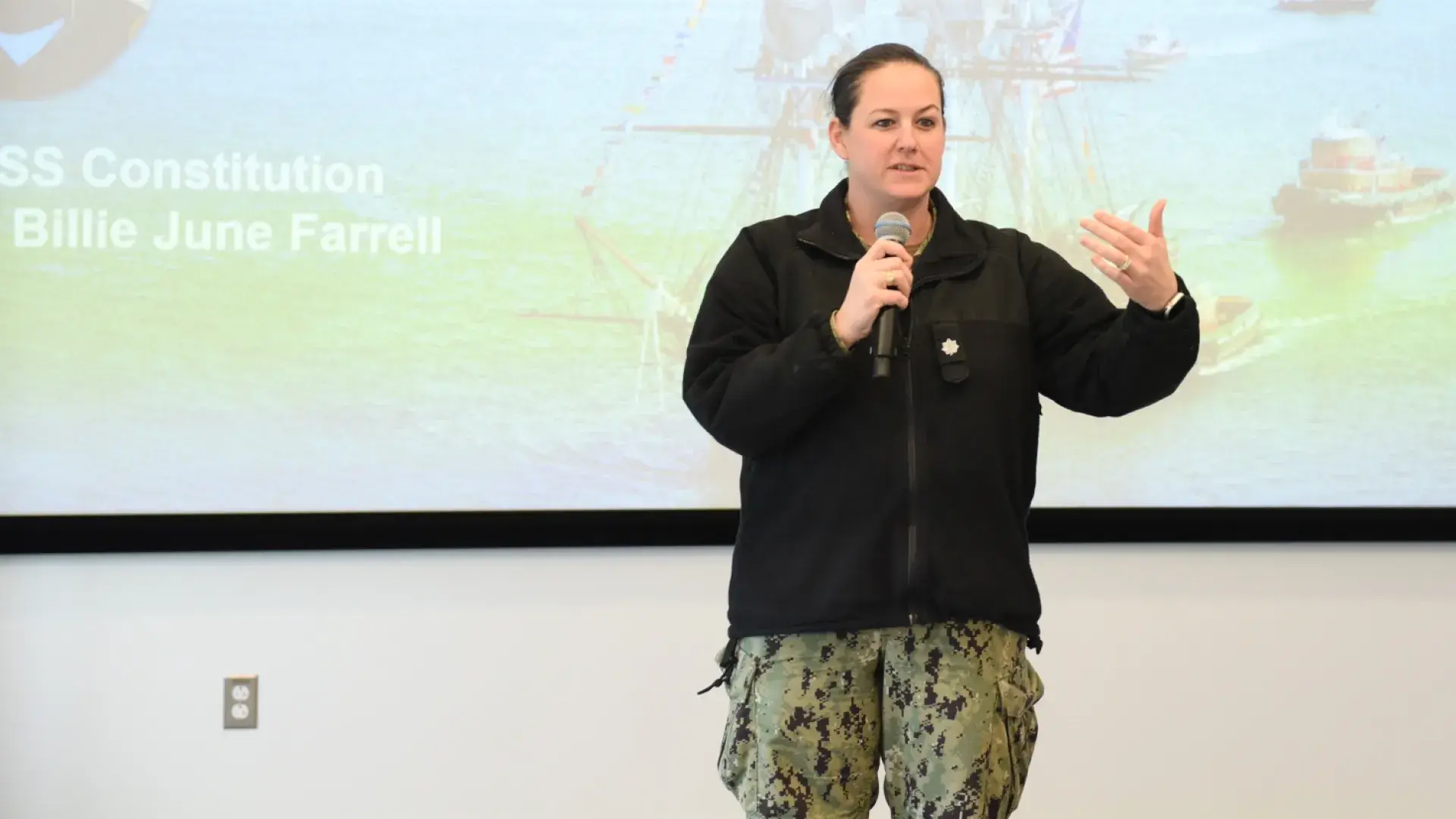 woman in green and black fatigue pants and black jacket speaks into a microphone