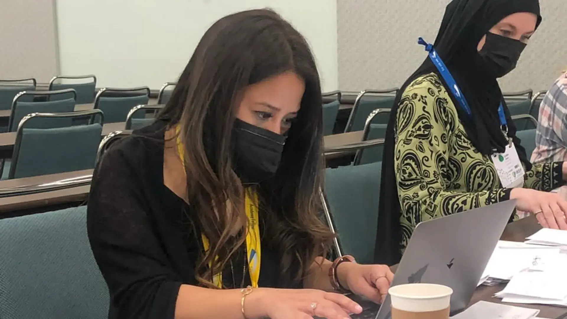 woman sits in chair at laptop next to another woman wearing a hijab