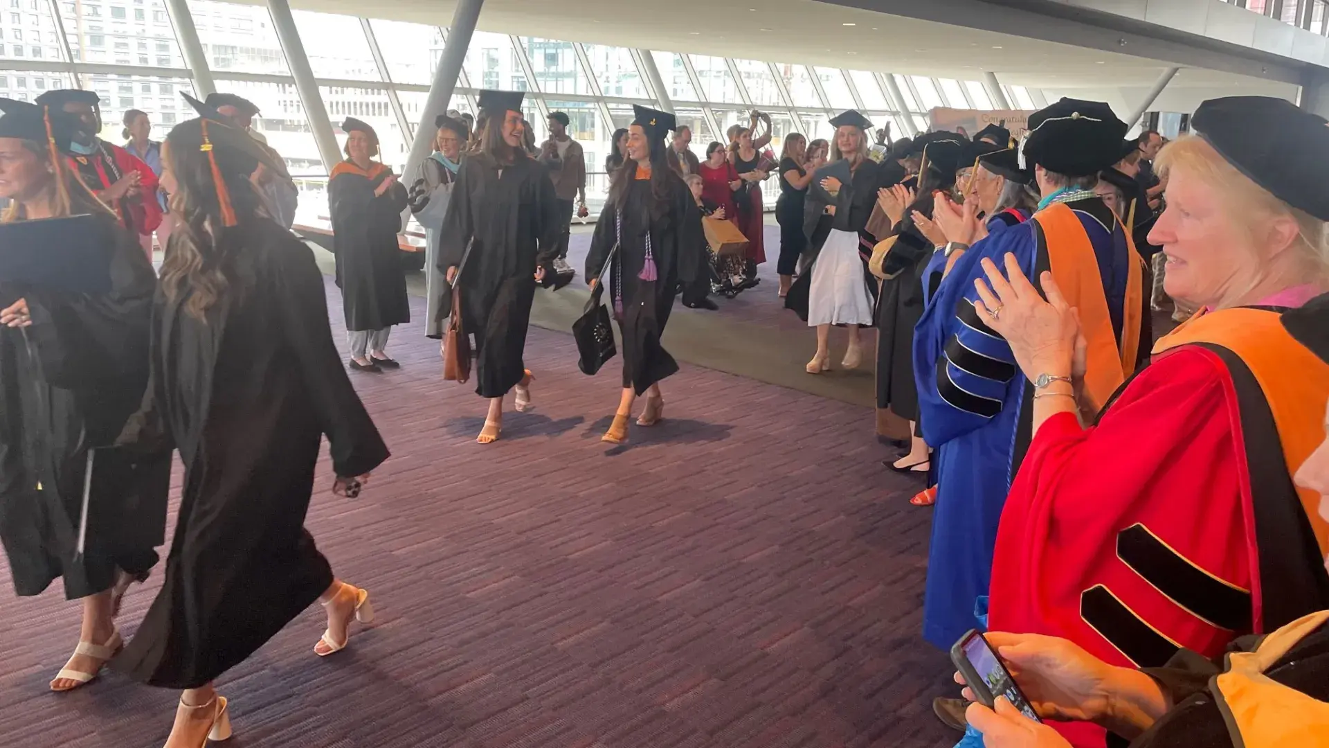 faculty in colorful graduation regalia clap as people wearing black caps and gowns walk in a line