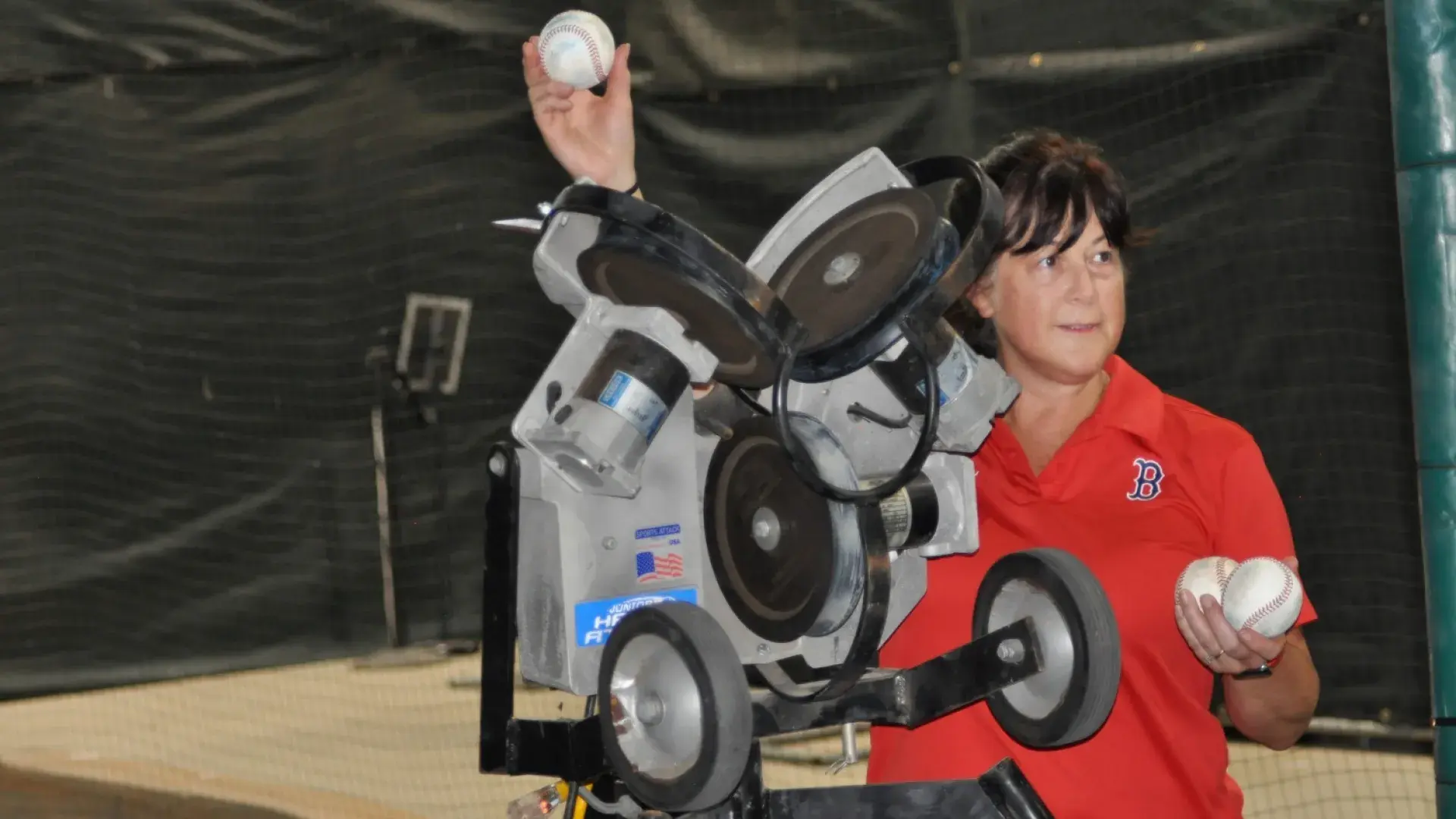 a woman in a red polo with a red sox logo B holds up a baseball next to a machine with wheels