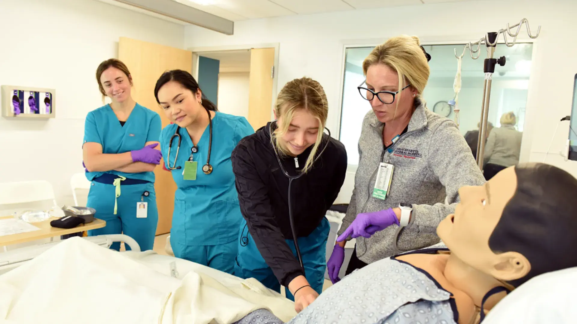 a woman in scrubs takes the pulse of a manikin in a bed while a woman in a MGH fleece looks on
