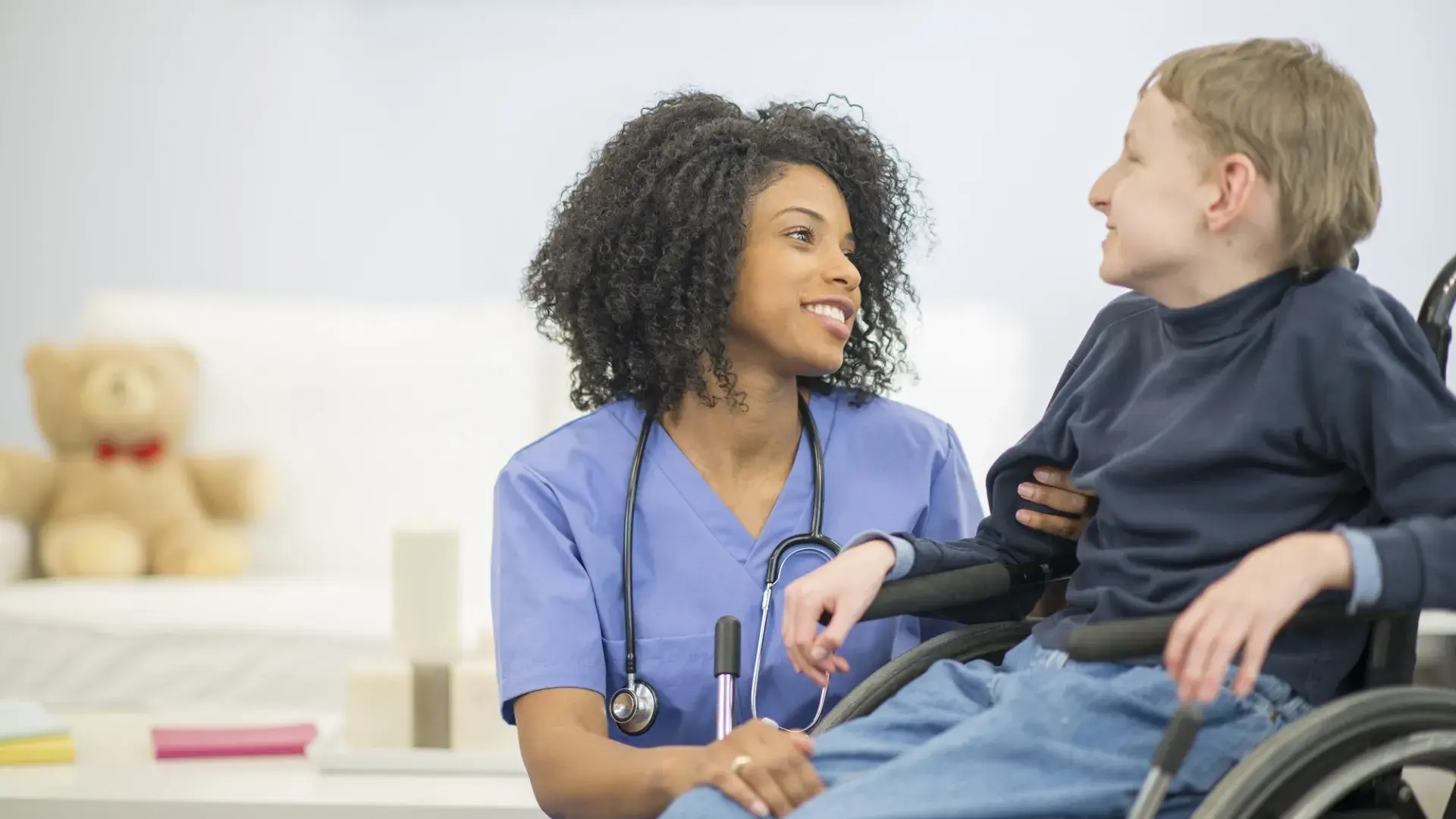 woman in blue scrubs crouches with a smile next to a small in stature person in wheelchair