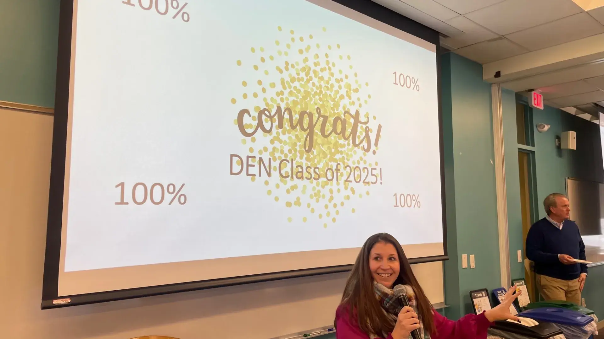 Faculty member delivering a congratulatory speech to MGH IHP DEN Class of 2025 celebrating 100% NCLEX first-time pass rate.