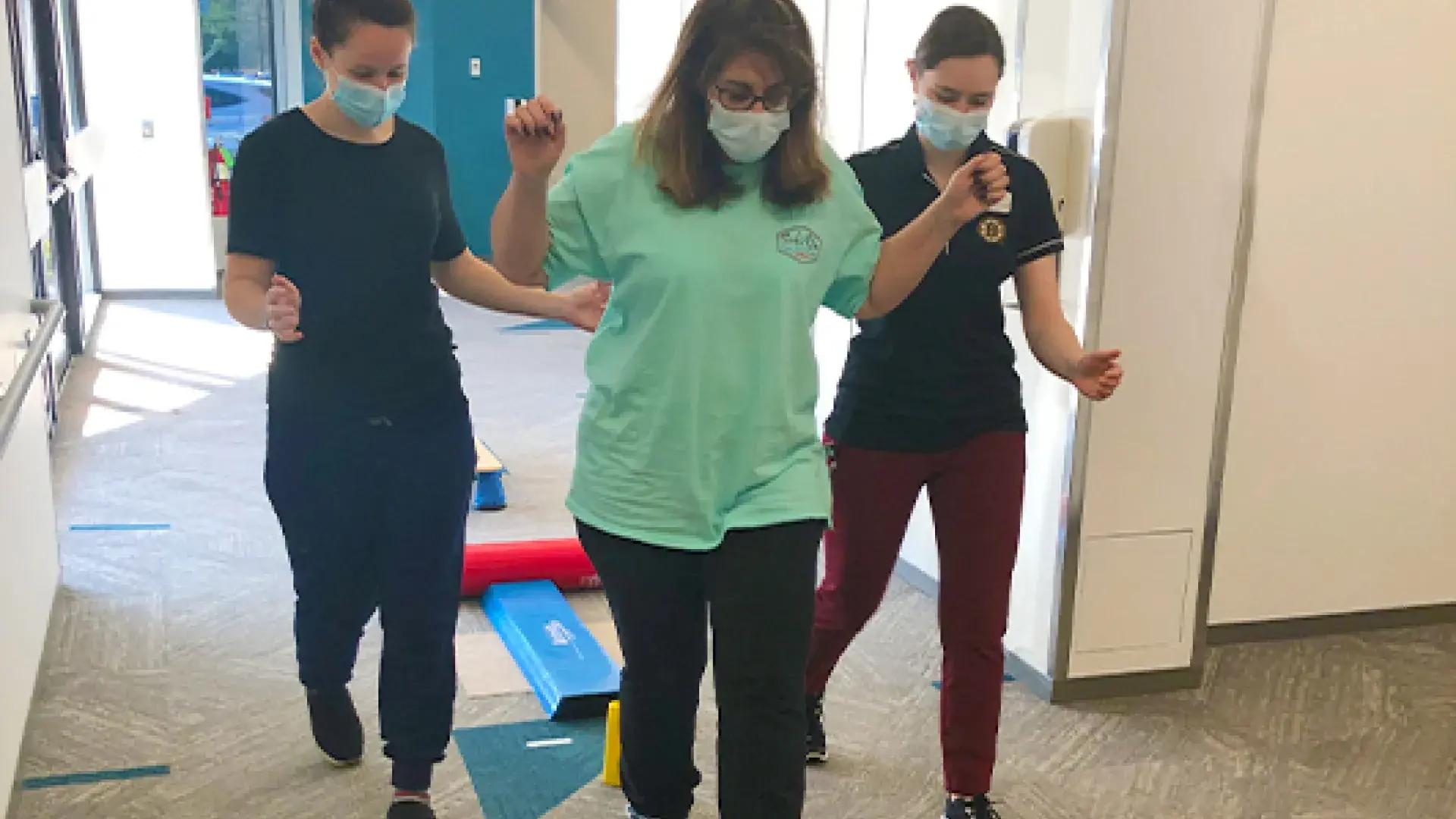 two women assist a third woman stepping over a low stick across two cones