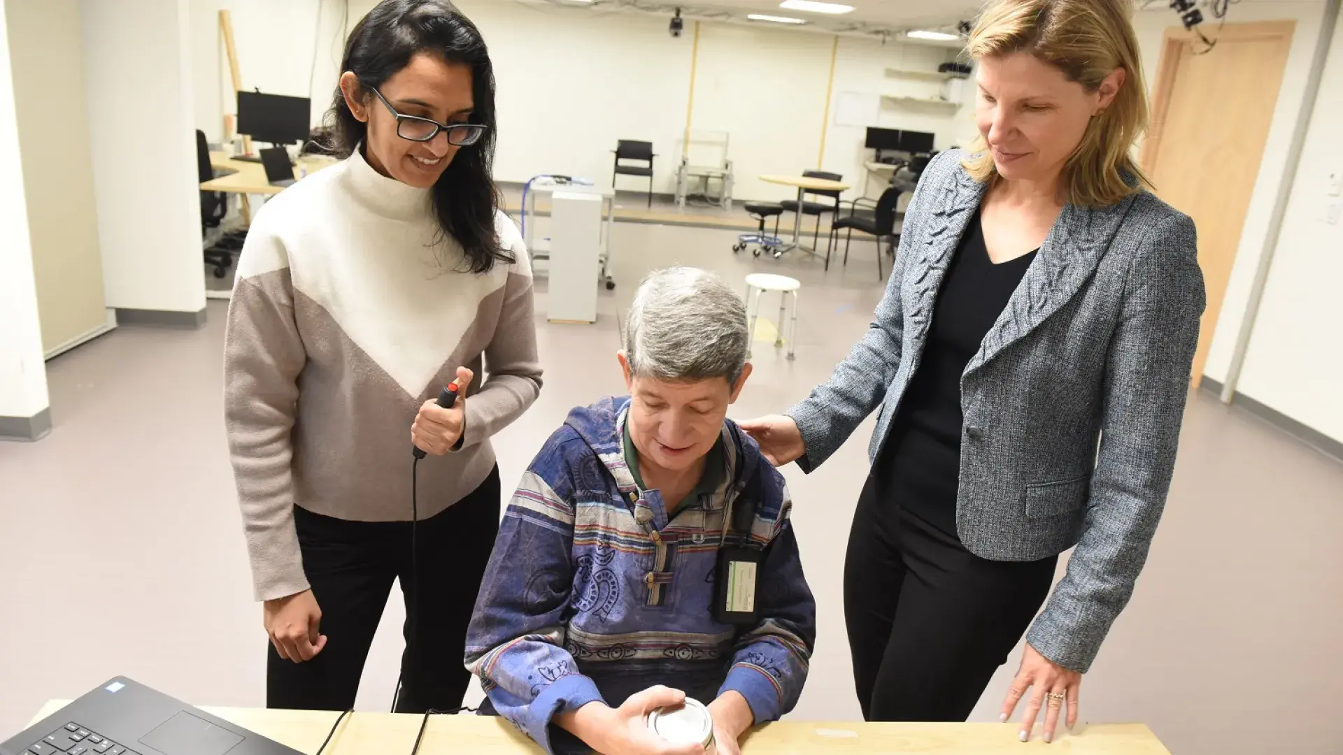 two women stand behind a person sitting at a desk one woman has her hand on the shoulder of the woman sitting down