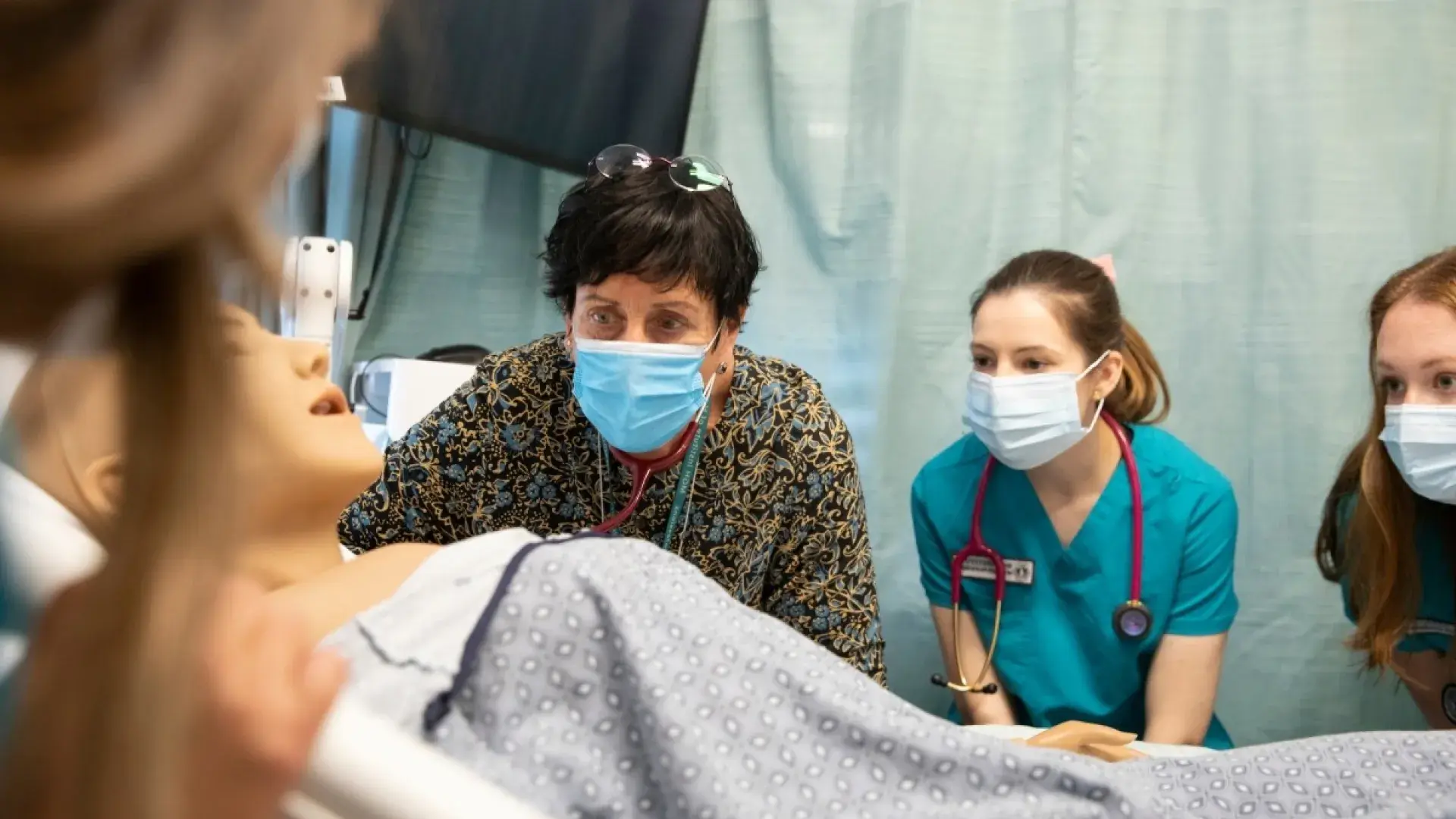 two women in scrubs and masks look at a manikin along with their teacher