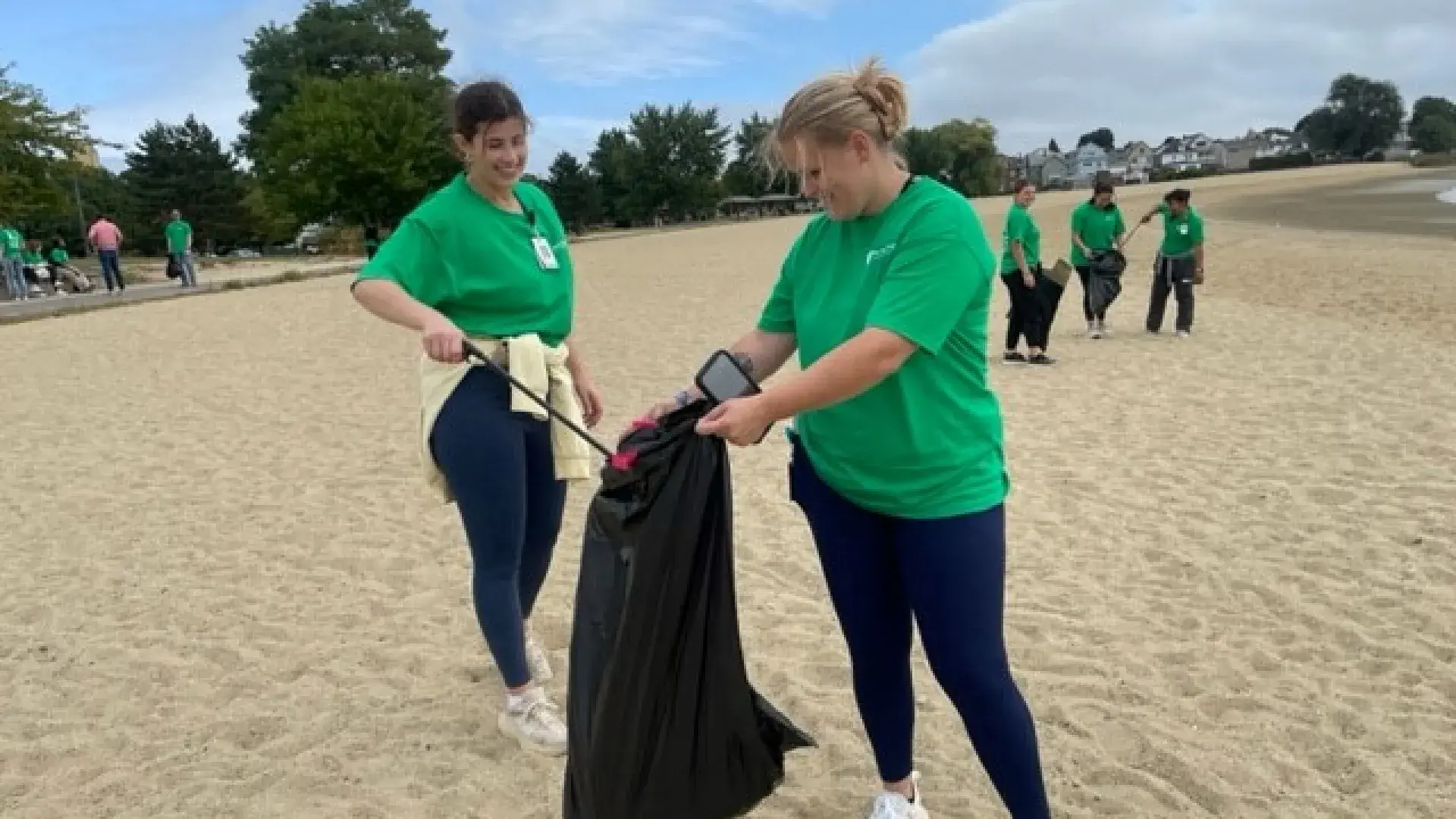 Two women on a beach with one holding a trash bag and then other using a tool to put trash in the bag