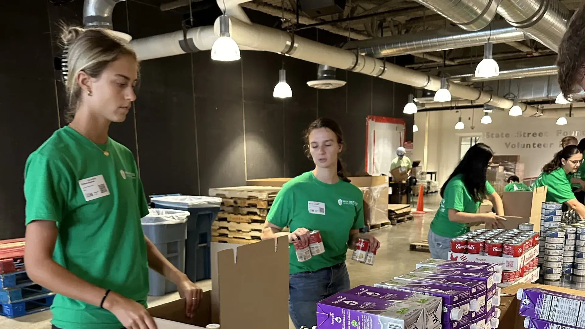 A woman moves a box down a conveyer belt to another woman holding cans of food while two other people further down the line add cans to boxes