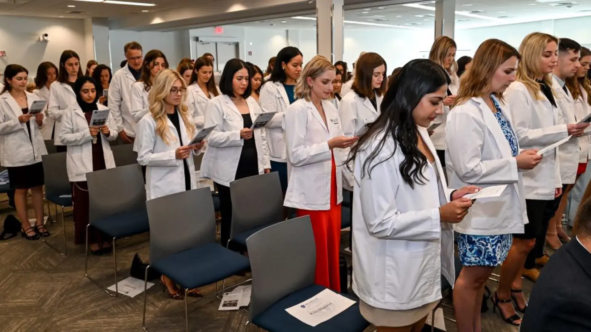 People in an audience, all wearing white coats, read from a piece of paper