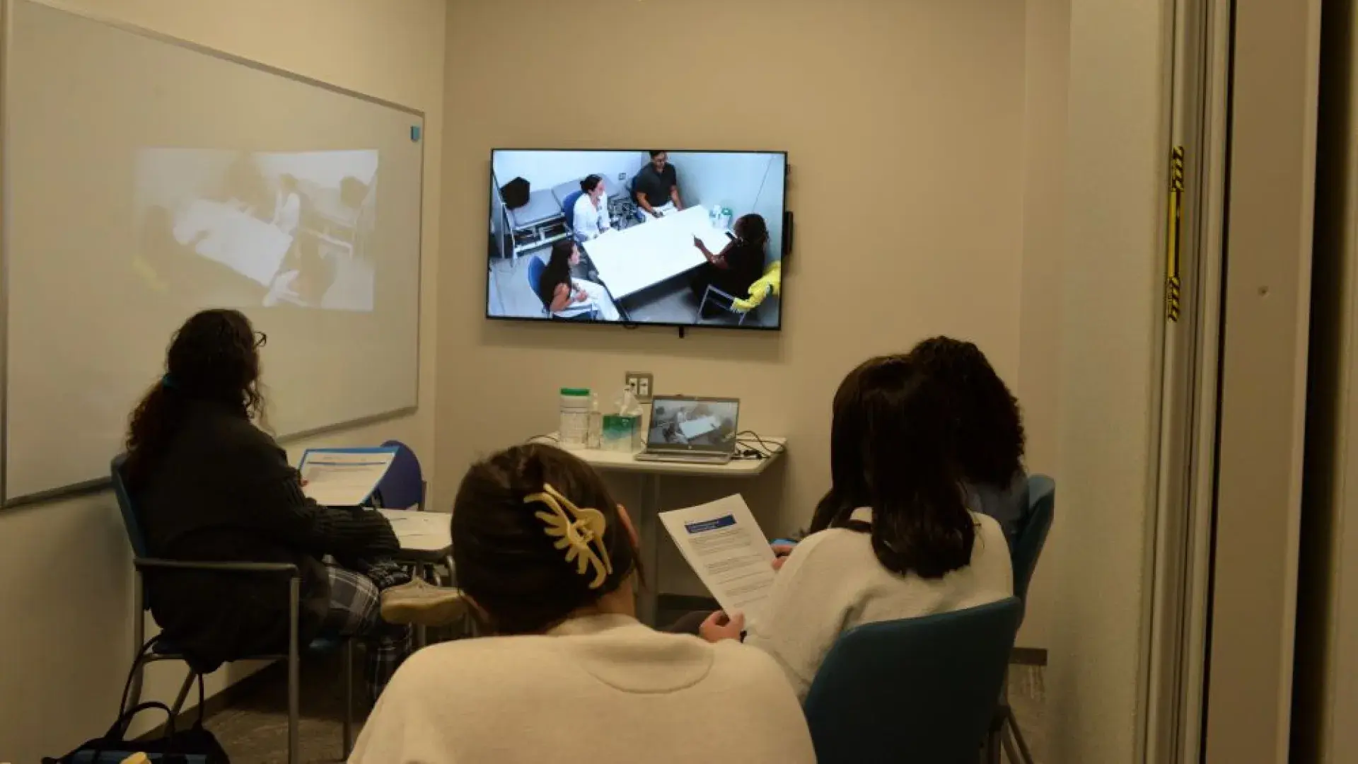 Four people sit in a room watching a screen that shows people sitting around a table