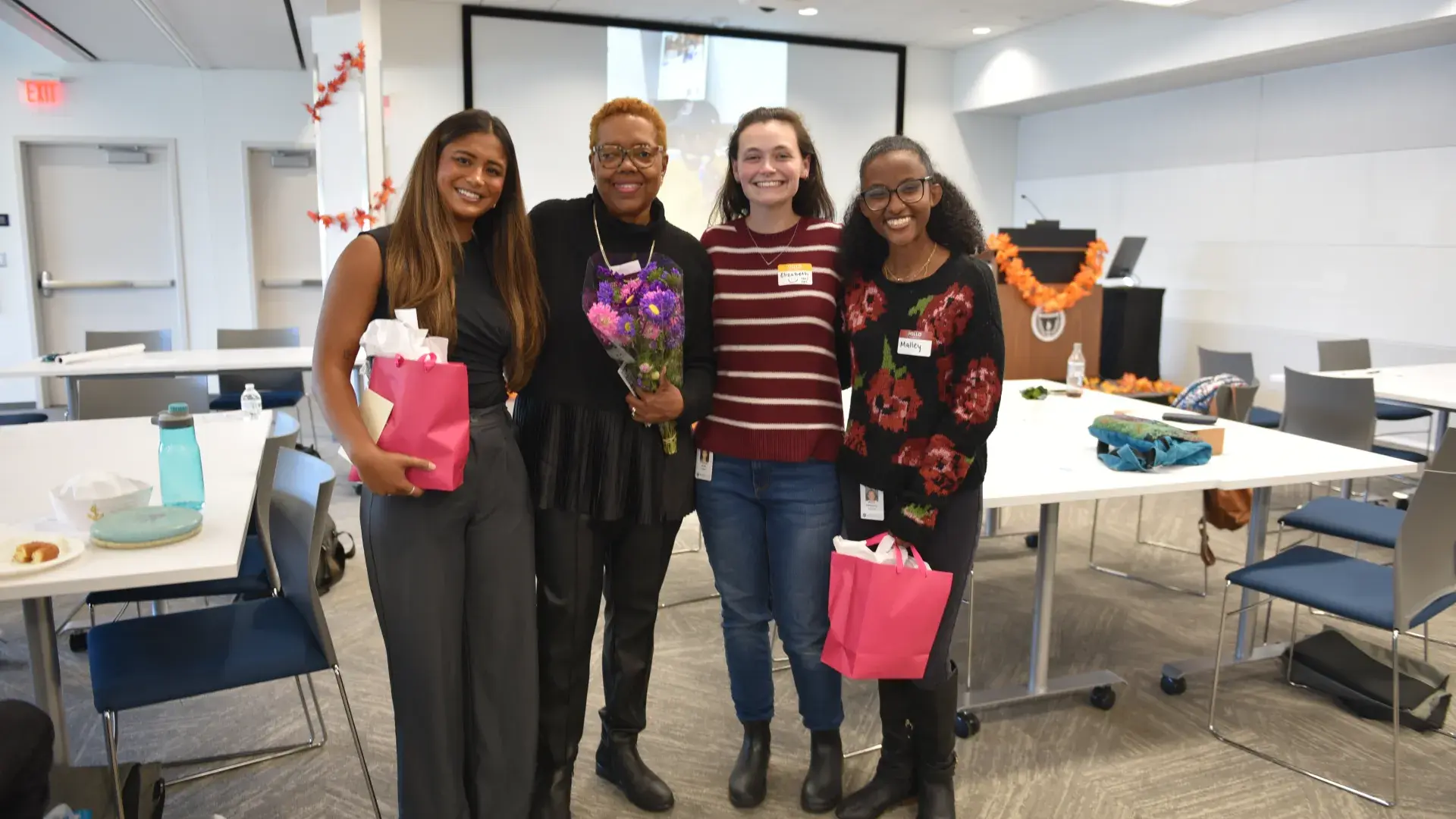 Four people pose with tables behind them and two of them are holding pink gift bags
