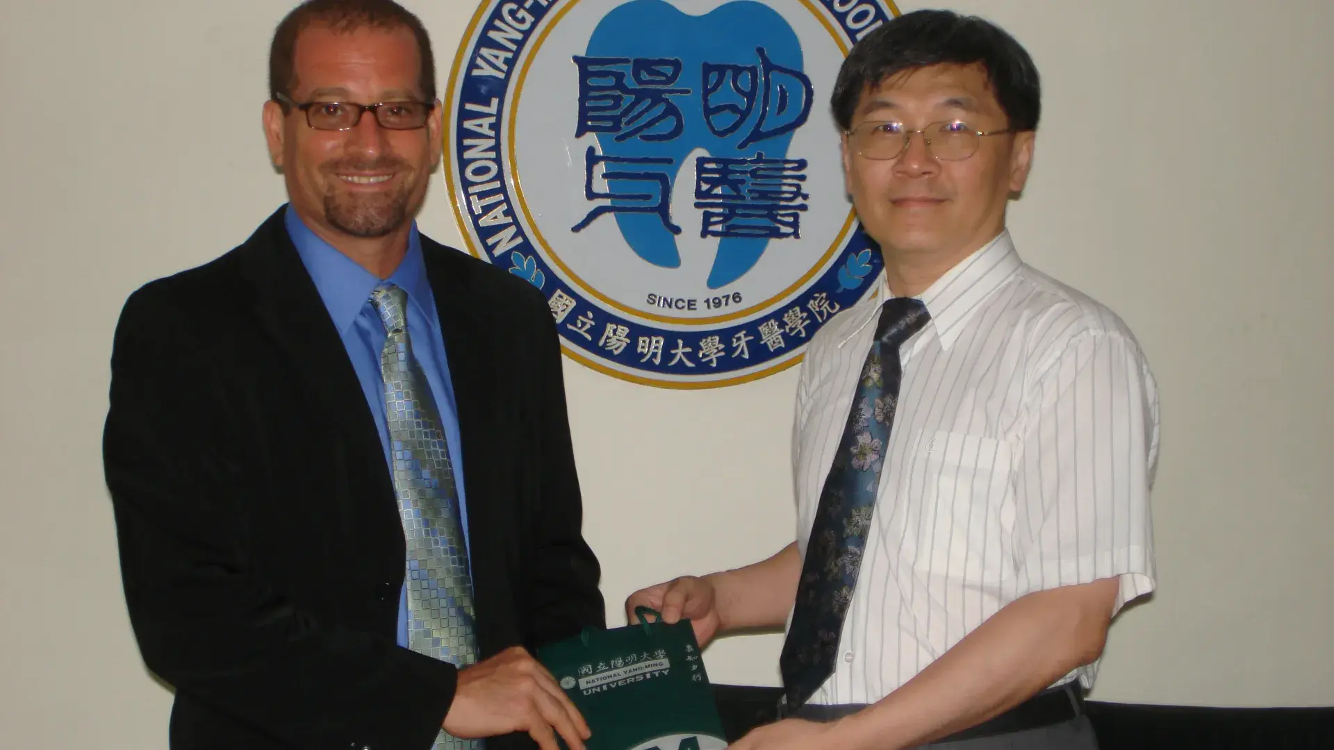 Two men stand in front of a school's logo 