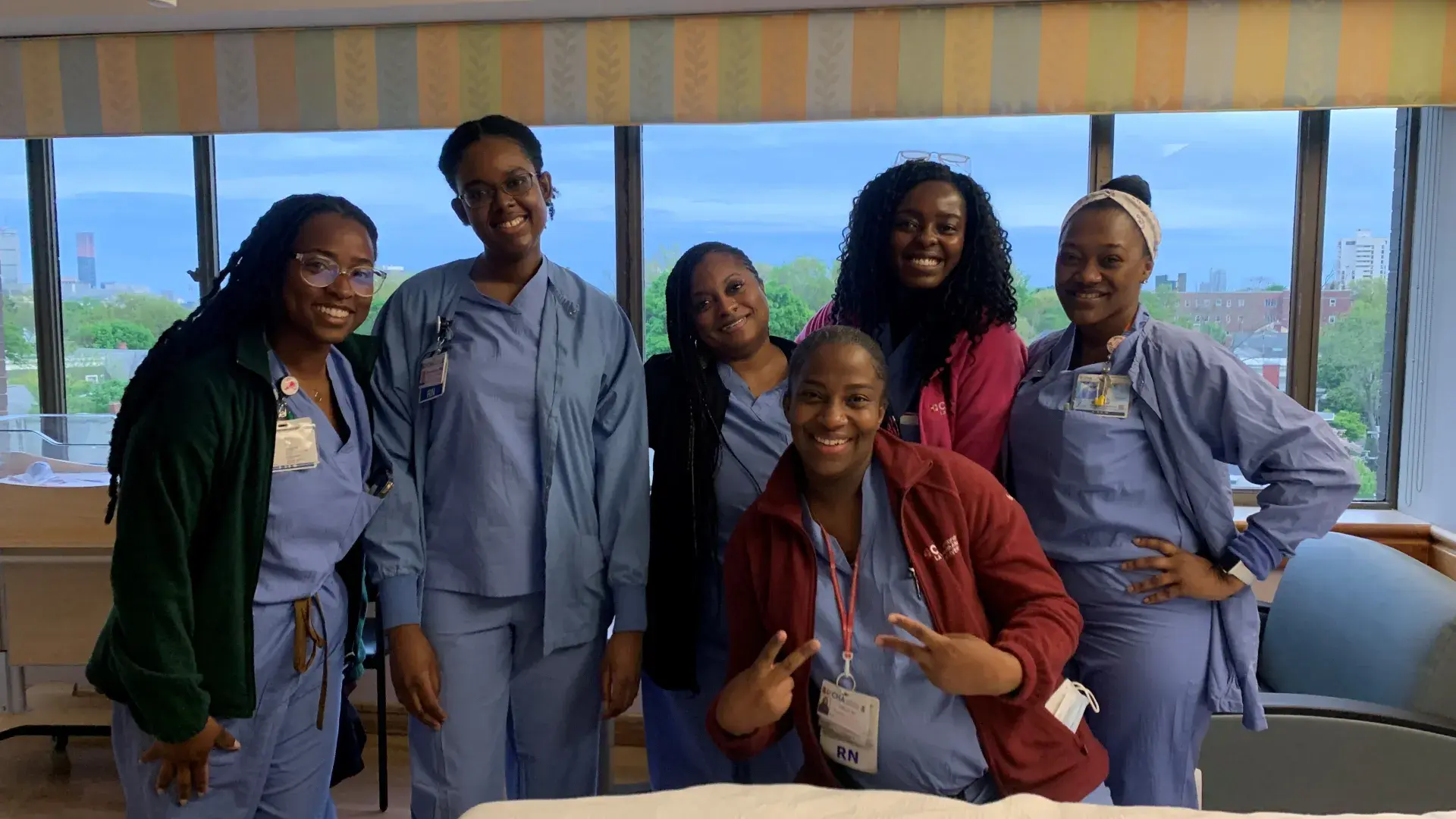 Six women in medical scrubs pose in front of a window