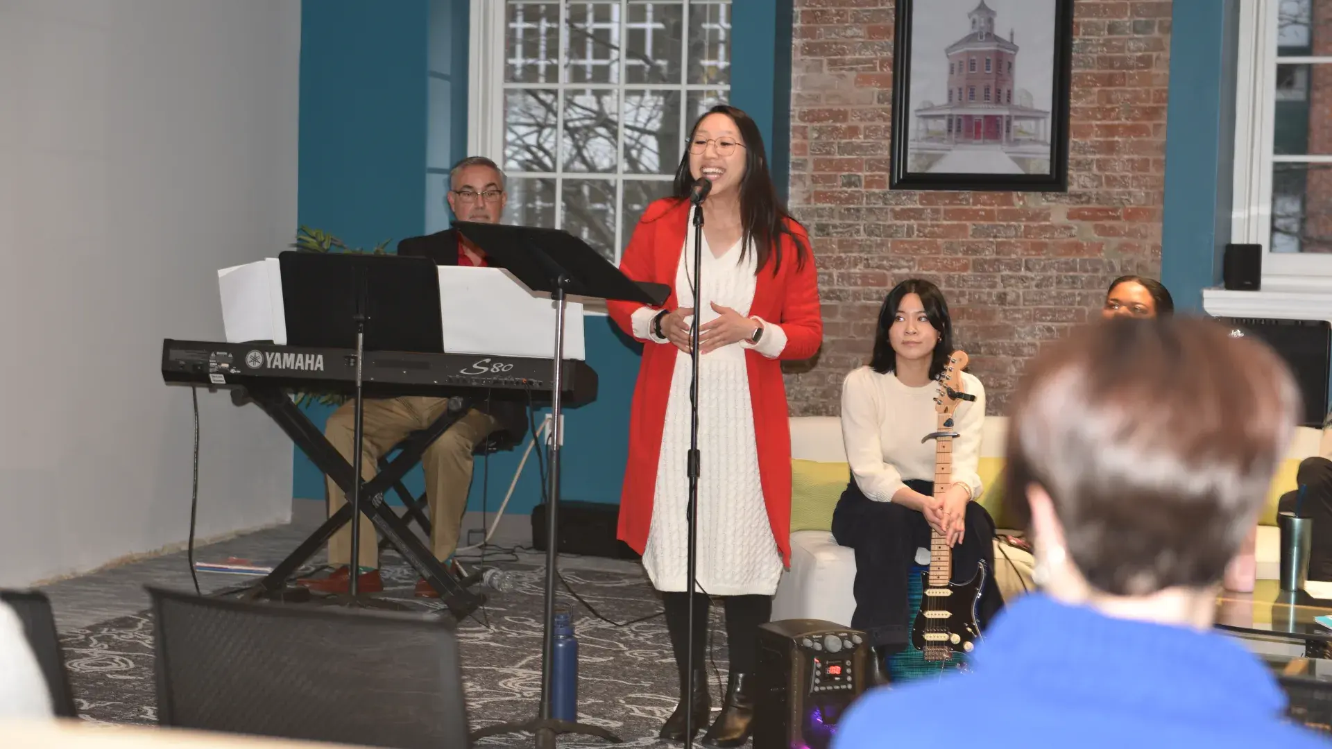 A woman sings into a microphone while a man plays the keyboard behind her and a woman holding a guitar sits on a couch. 
