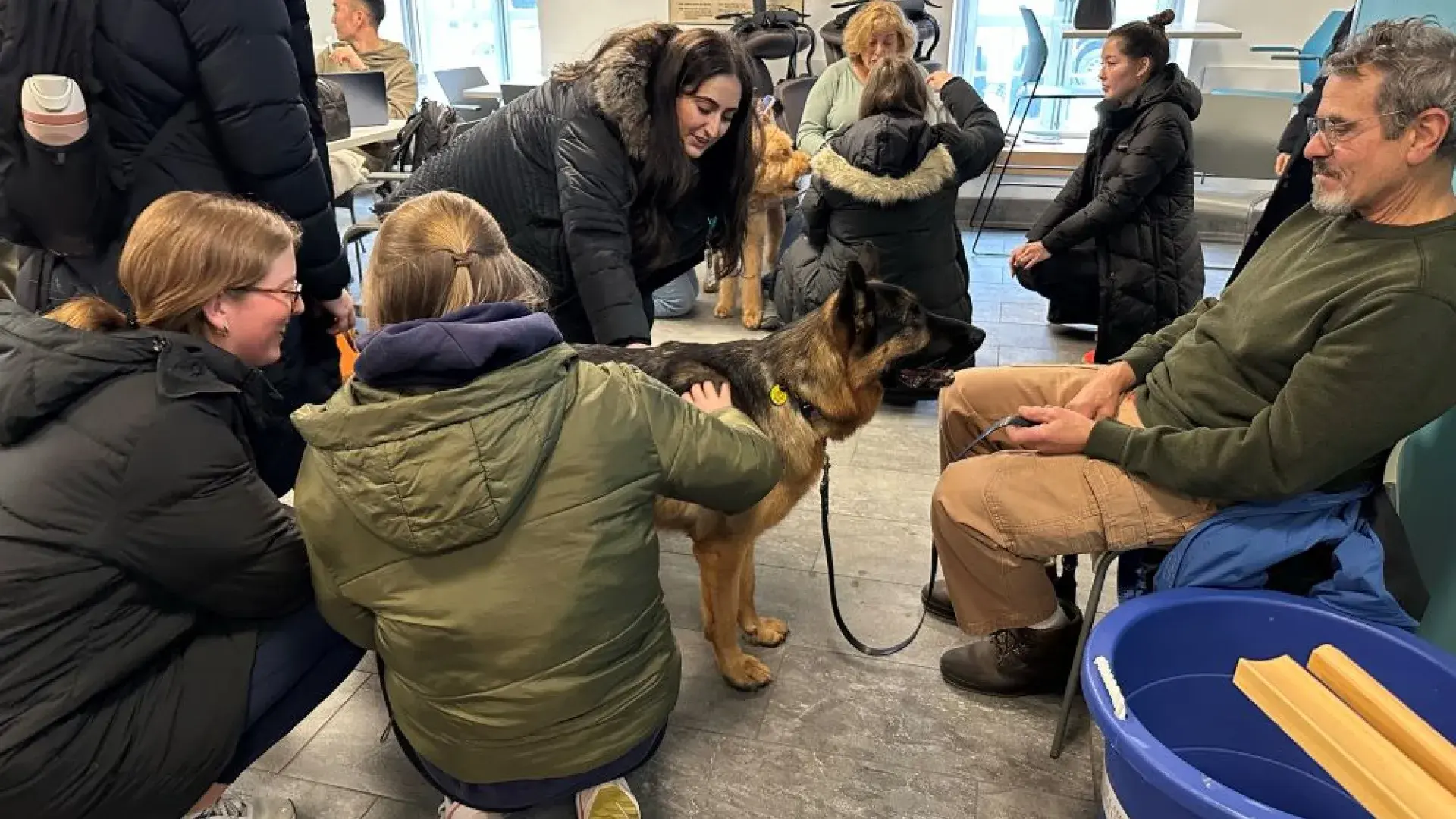 Three people pet a dog while a man holding the leash sits in a chair
