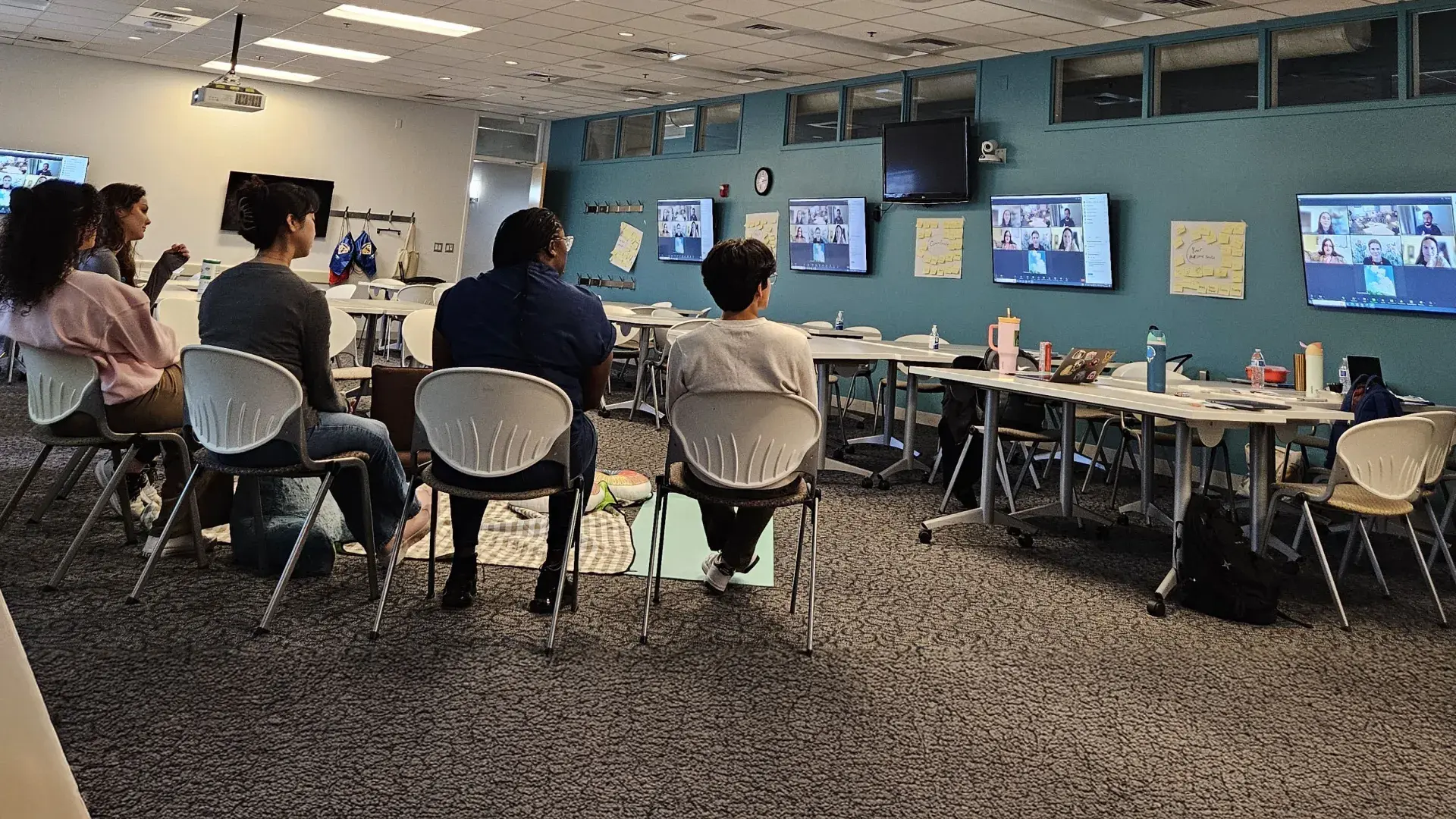 Six people sit in a semi circle watching screens with a virtual meeting