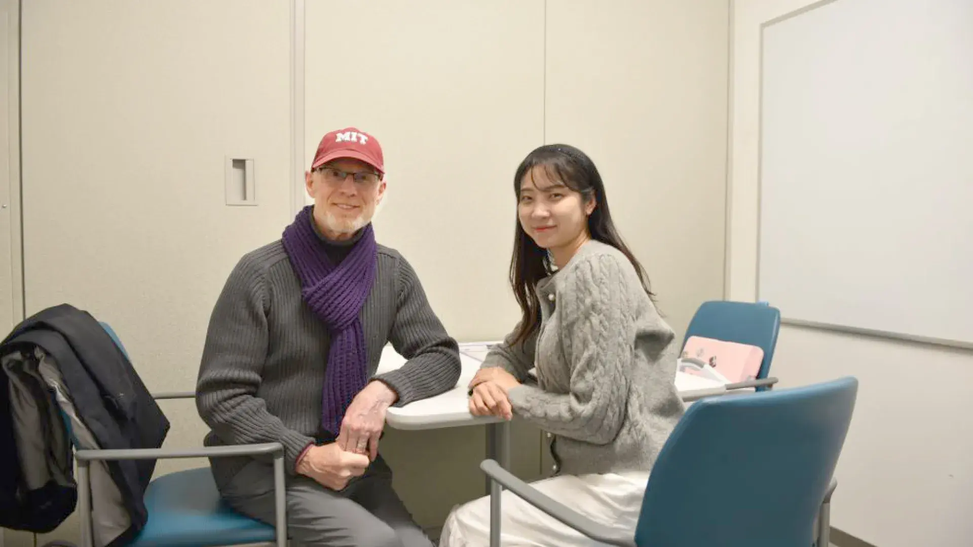 A man in a red baseball cap sits at a table facing the camera while a woman sits next to him facing the camera