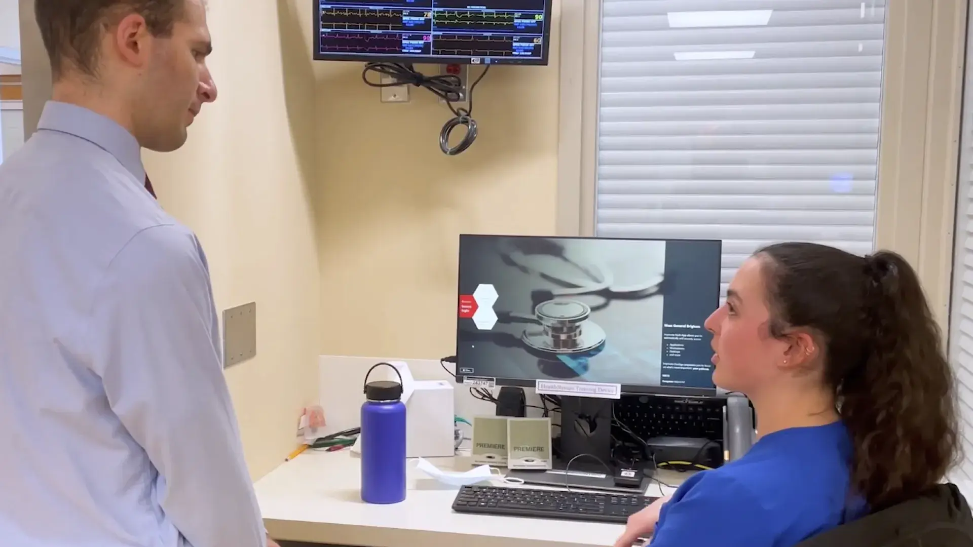 man in dress shirt speaks to a woman in scrubs at a computer