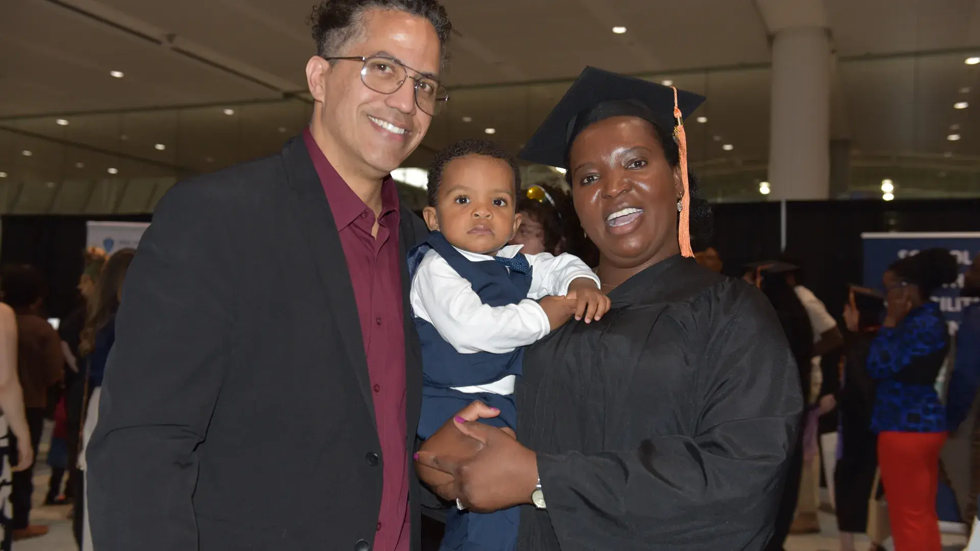 A man and a woman, who is wearing a graduation robe, pose with a baby